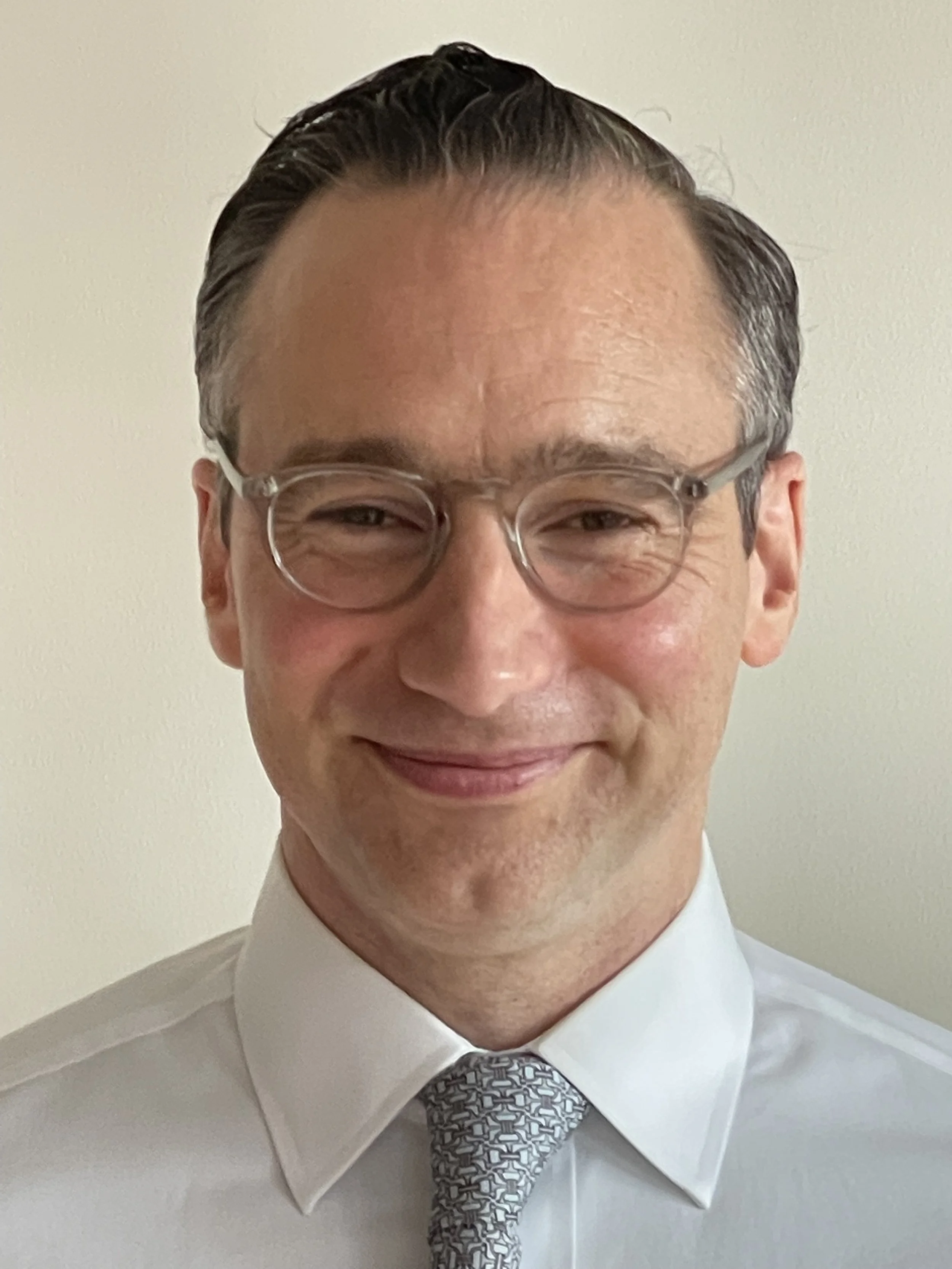 A man with slicked-back hair, glasses, and a white dress shirt with a patterned tie, smiling against a plain light background.