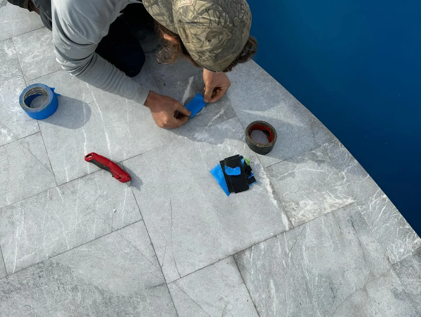A person with a camo cap and gray long-sleeve shirt kneels on a tiled floor, applying blue painter's tape near a blue wall, with tools including a utility knife, rolls of tape, and a small tool pouch scattered around.