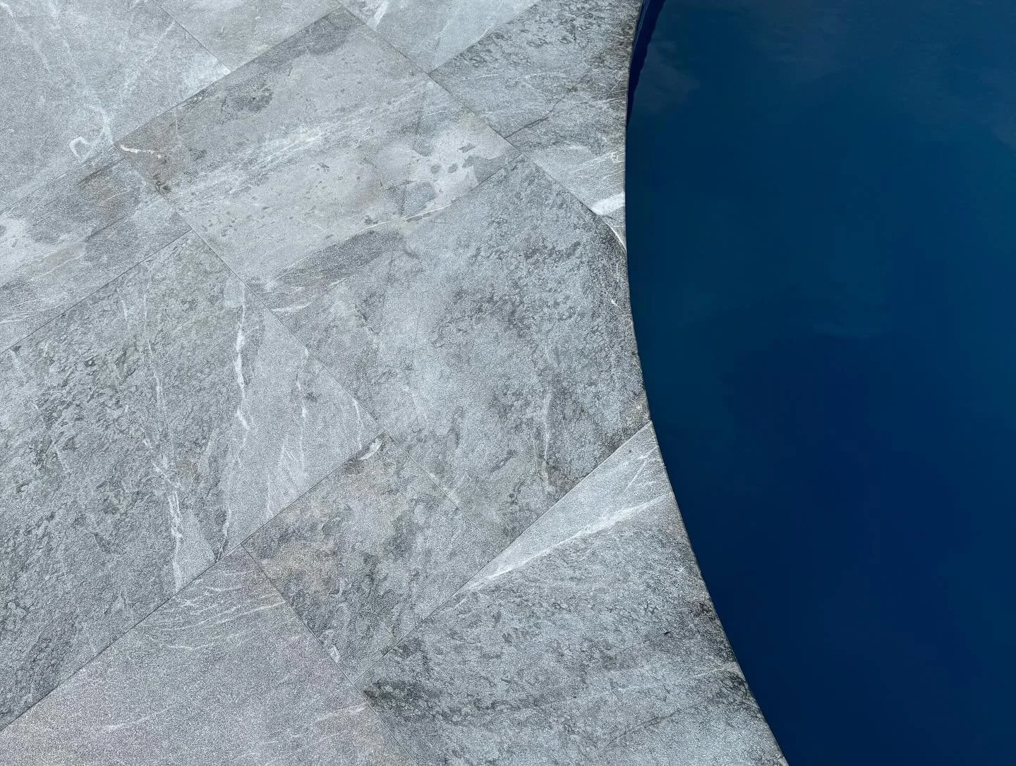Close-up of a tiled gray stone floor next to a blue swimming pool with curved edge.