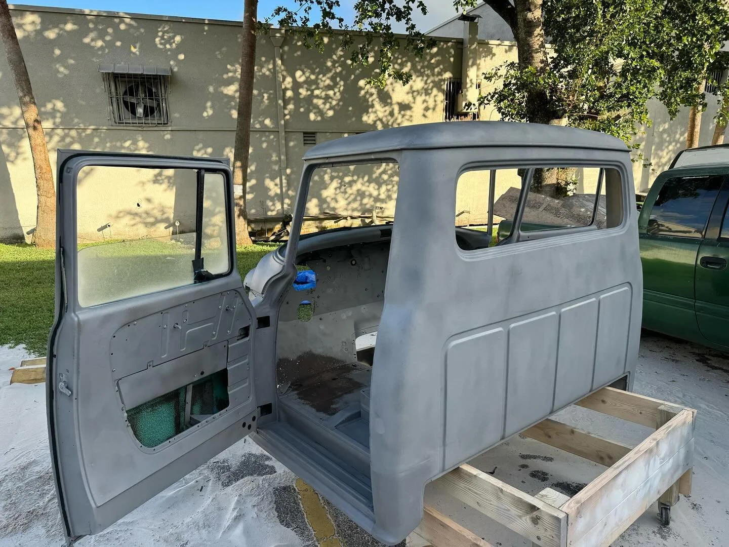 An unfinished vintage vehicle body, possibly a classic car, mounted on a wooden frame outdoors with trees and a building in the background.