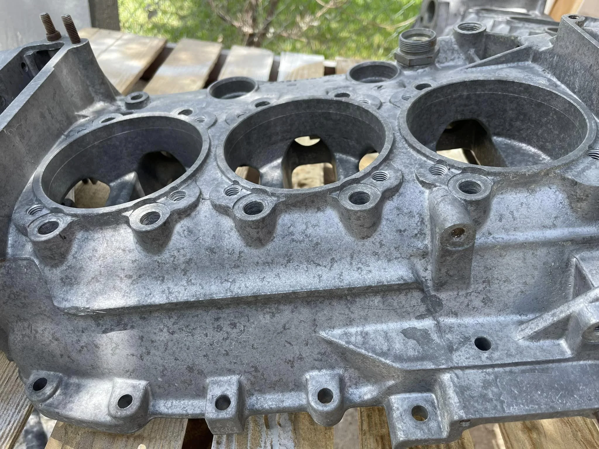 A metallic engine block with three large cylinders, placed outdoors on wooden pallets.