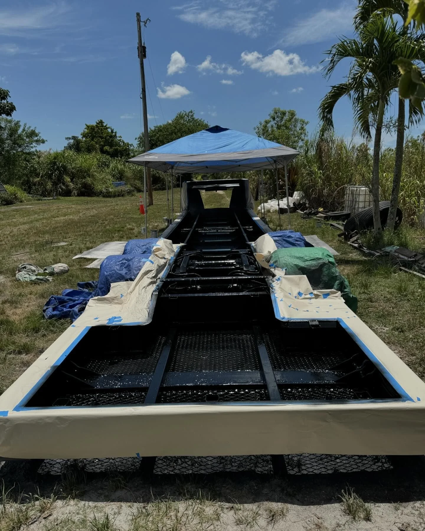 A boat under construction on a grassy area outdoors, with the hull painted black and covered with protective paper and tape, set up under a blue canopy. There are trees and a utility pole in the background.
