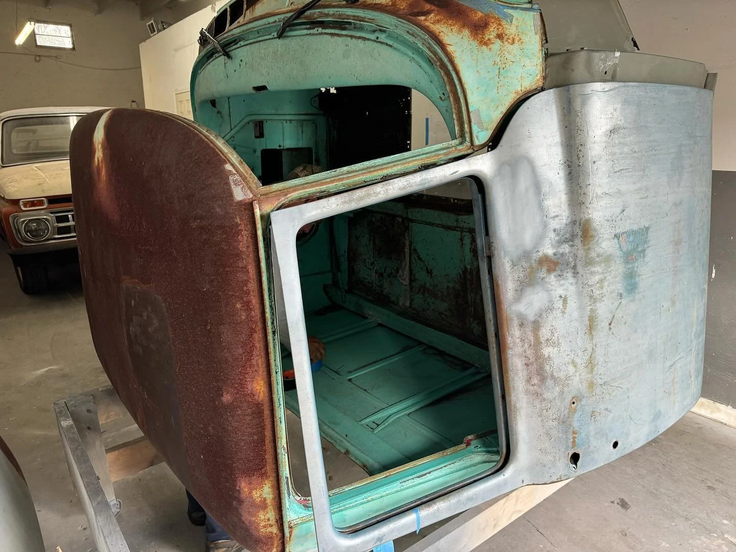 A vintage pickup truck in the process of restoration, showing a rusted hood, and a weathered cab with a partly stripped interior, inside a garage with another classic vehicle in the background.