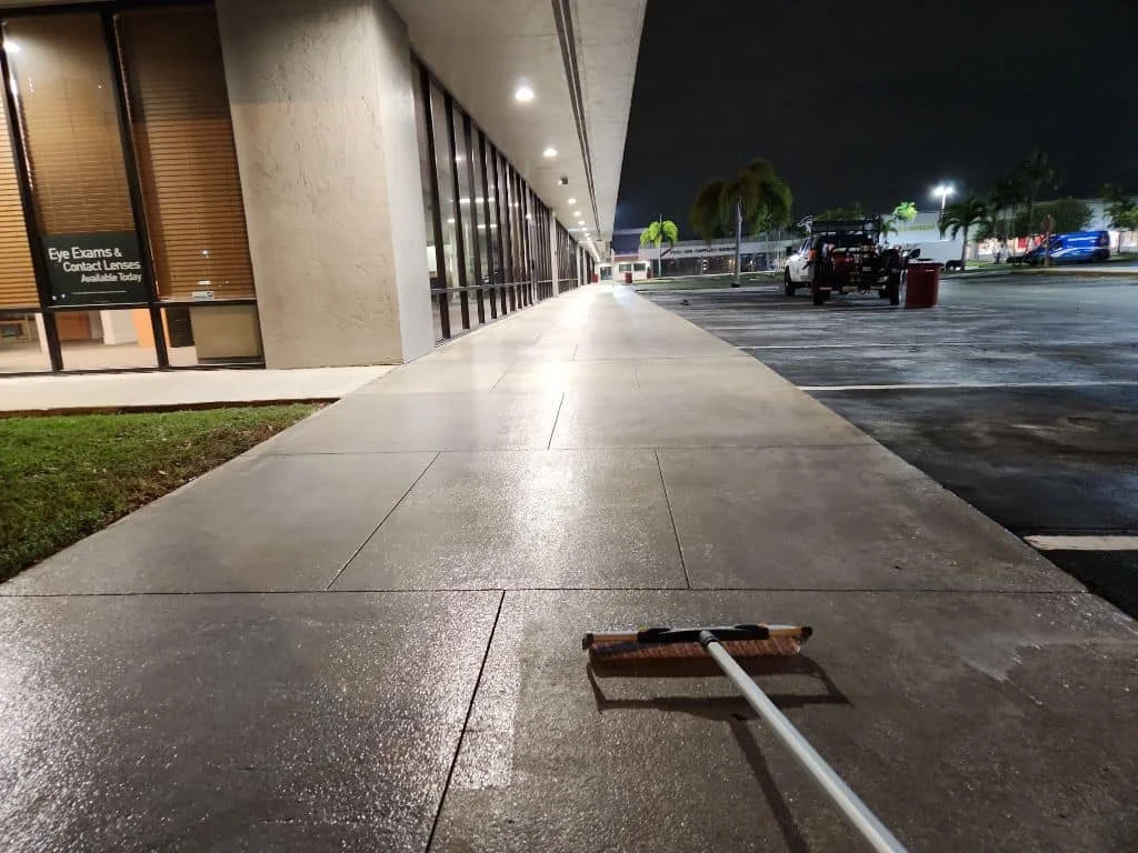 Empty sidewalk in front of a building at night, with bright lights and parked cars in the background. A push broom is lying on the sidewalk.