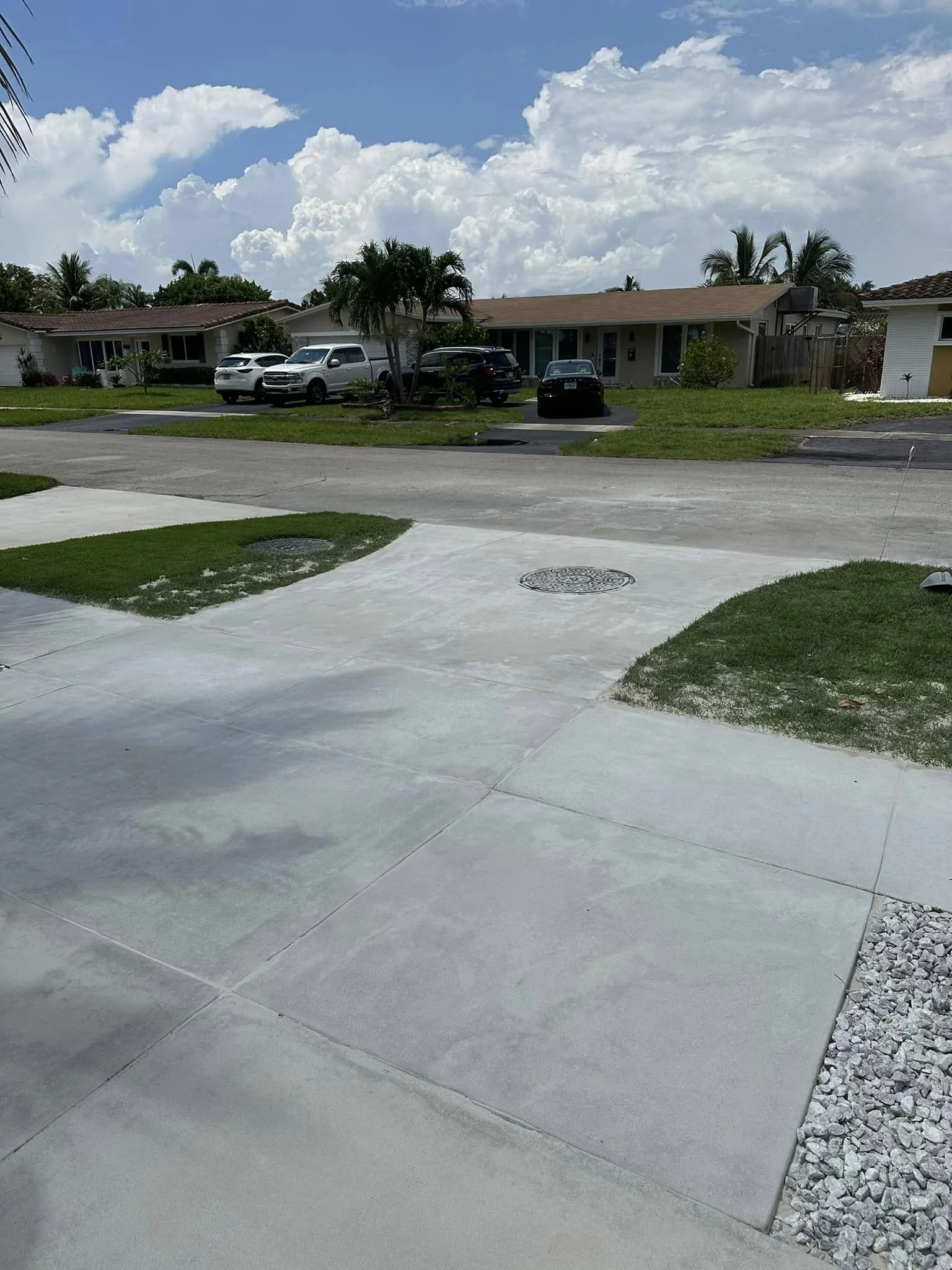 View of a suburban neighborhood with houses, palm trees, parked cars, a partly cloudy sky, and a concrete driveway in the foreground.