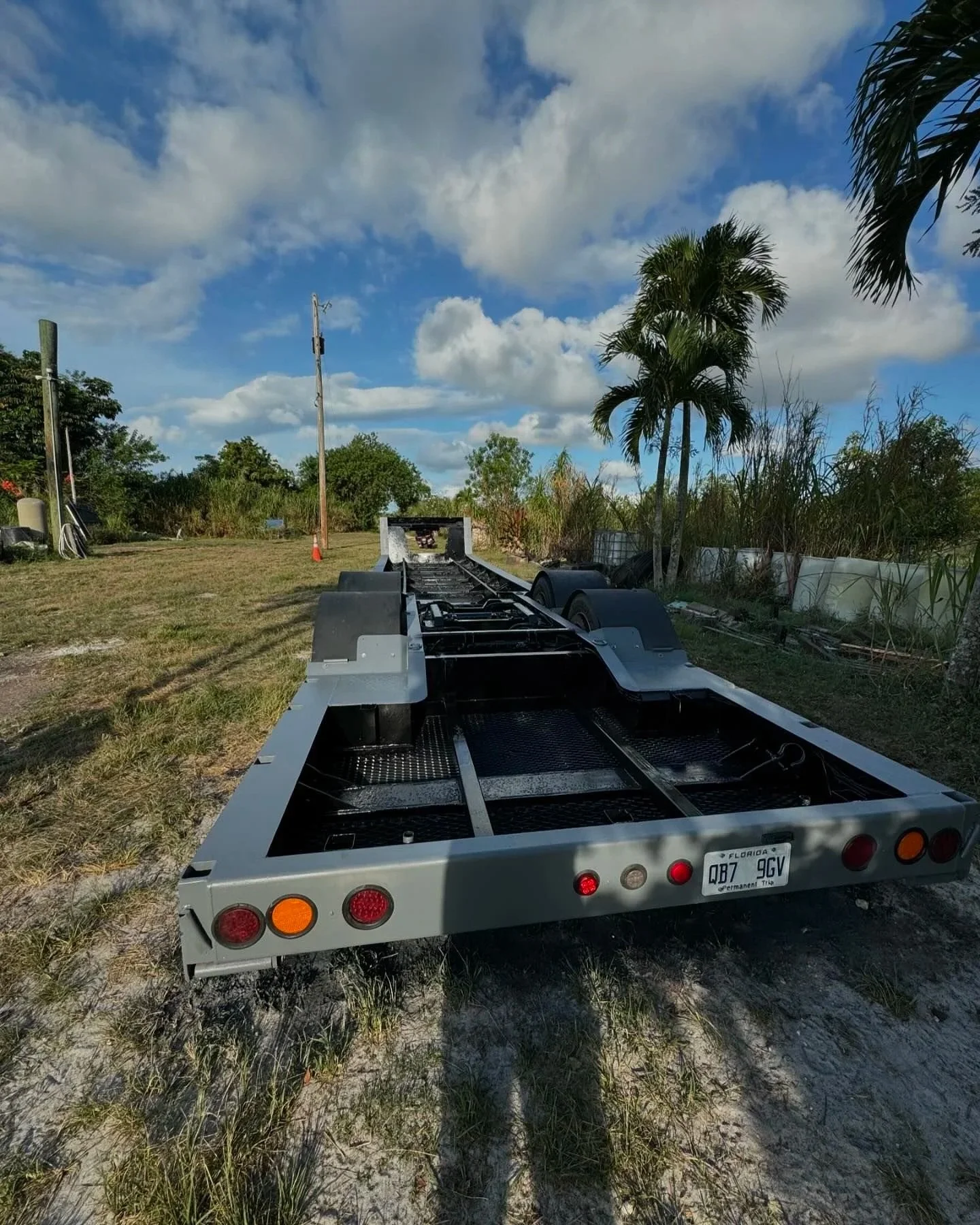 Empty flatbed trailer in a grassy outdoor area with trees and utility poles, under a partly cloudy sky. sandblasting and painting