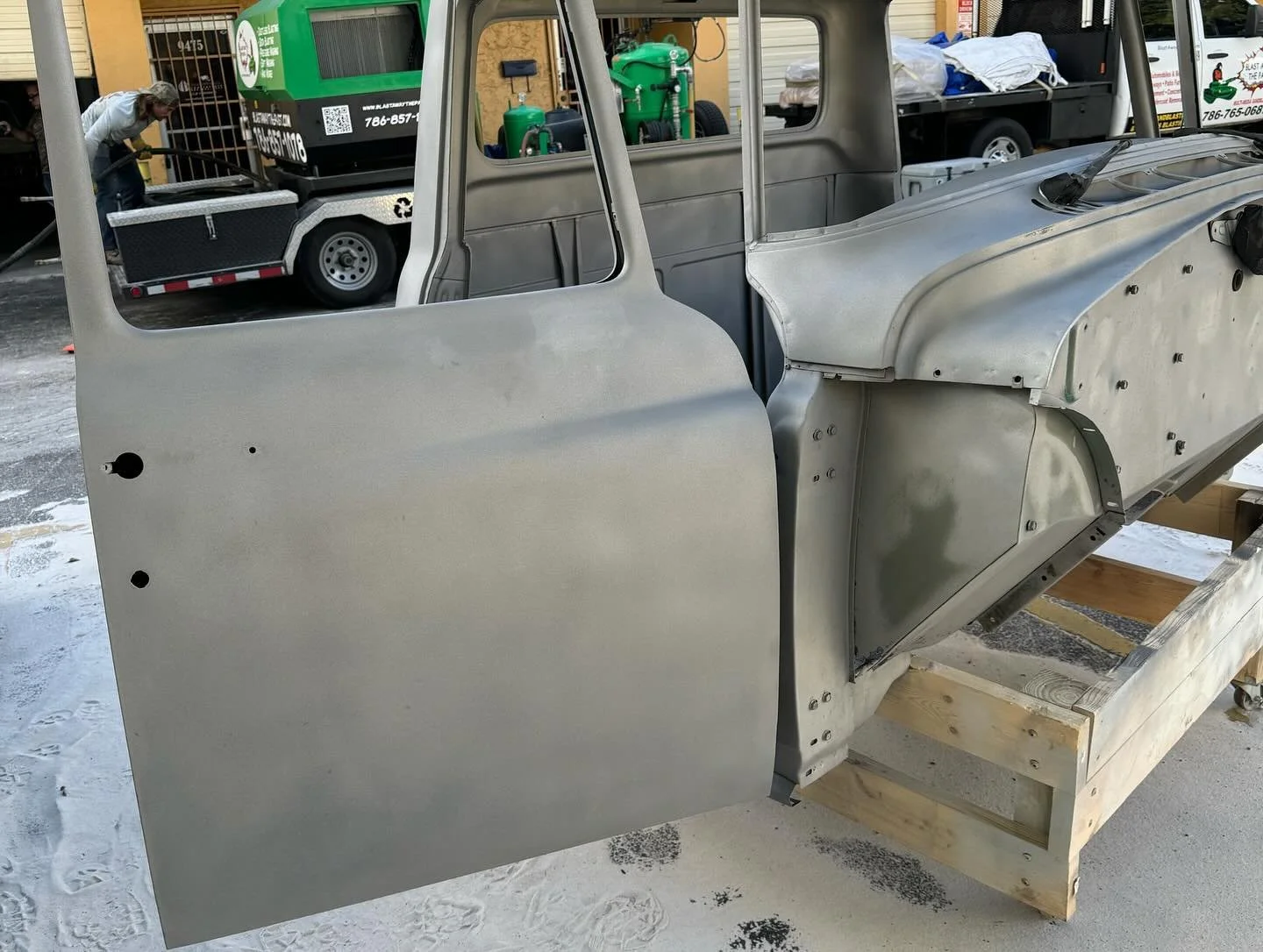 Unfinished metal vehicle body or frame on wooden support outside, with industrial equipment and a person working in background.