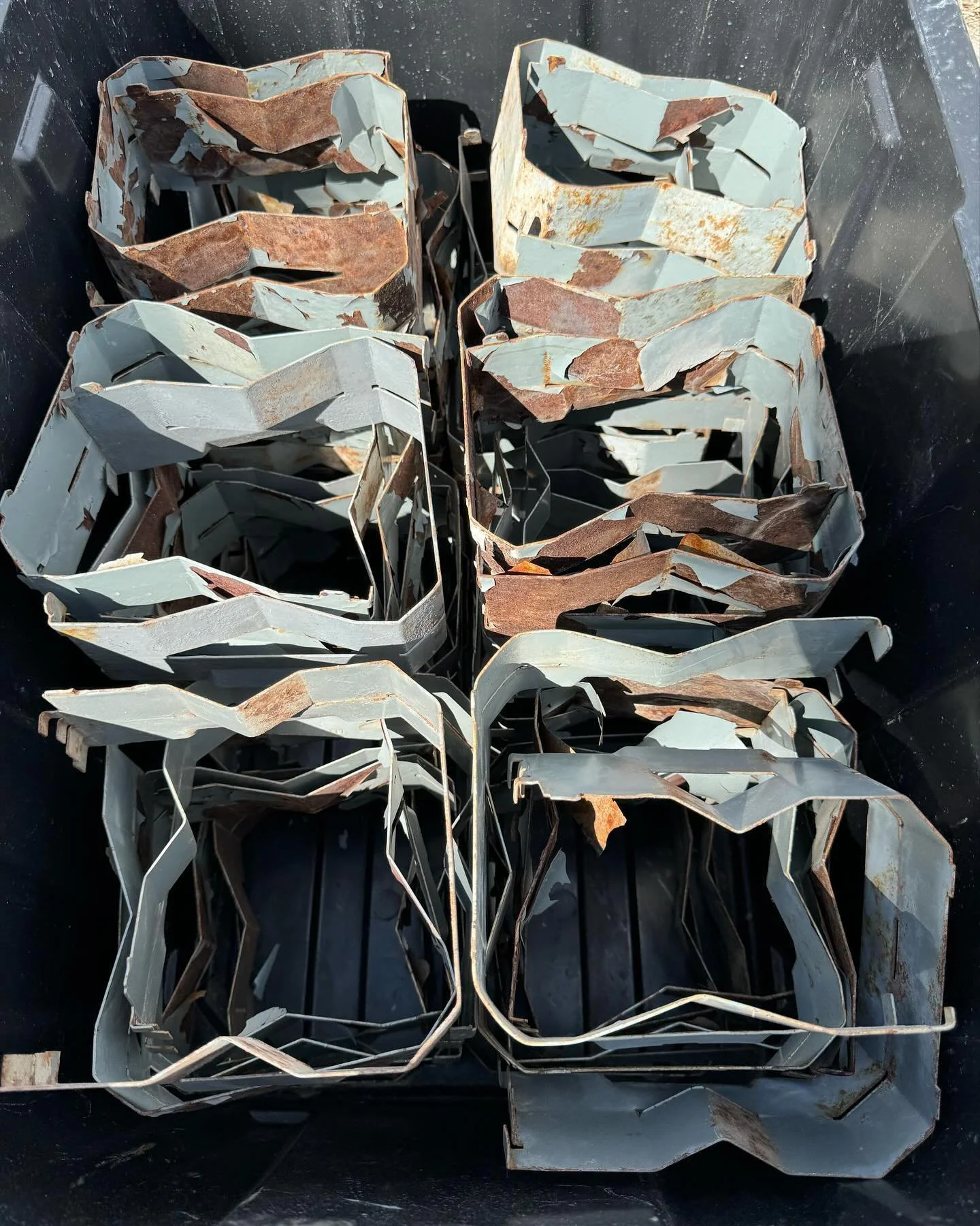 A collection of rusted and bent metal pieces organized in black containers, with some painted light blue and others showing rust, arranged in rows.