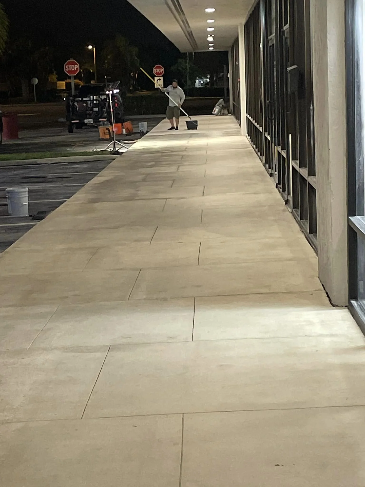 A person cleaning or mopping the sidewalk outside a building at night, with stop signs visible in the background.