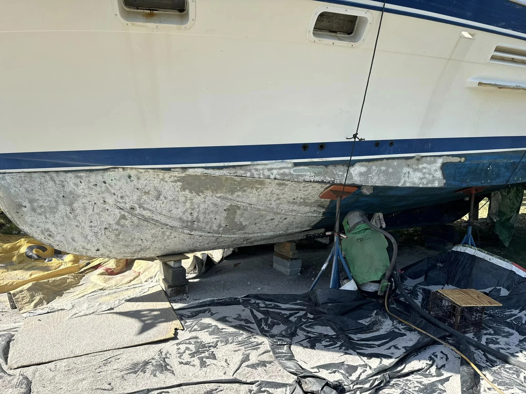 The hull of a boat elevated on supports with visible algae and grime, surrounded by waterproof sheets, a green vacuum, a crate, and other equipment.