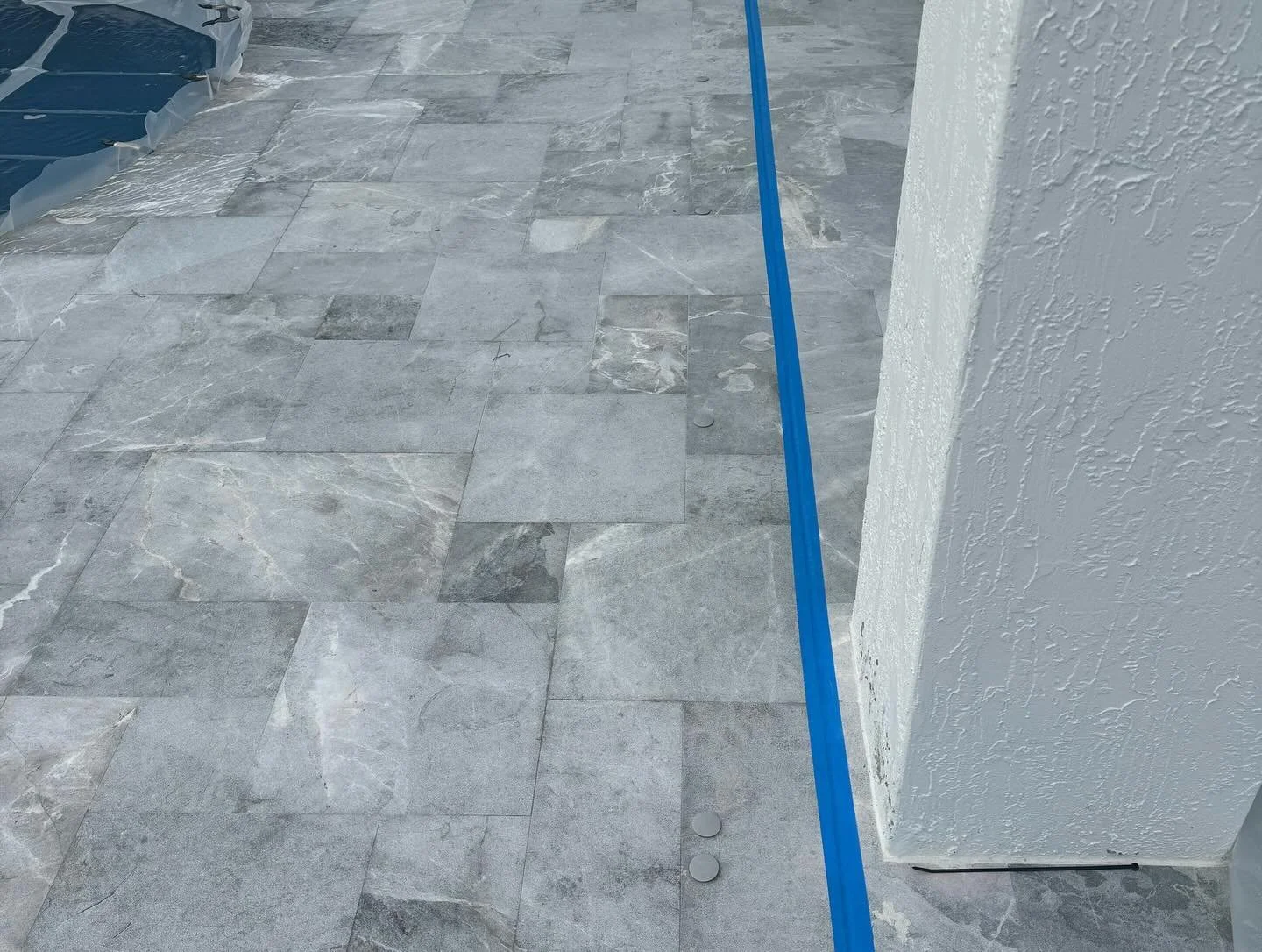Gray pool deck marble flooring with a white textured wall on the right, a blue measuring tape or straight edge, and a blue plastic sheet or cover partially visible on the top left corner. Sandblasted to remove failing sealer.