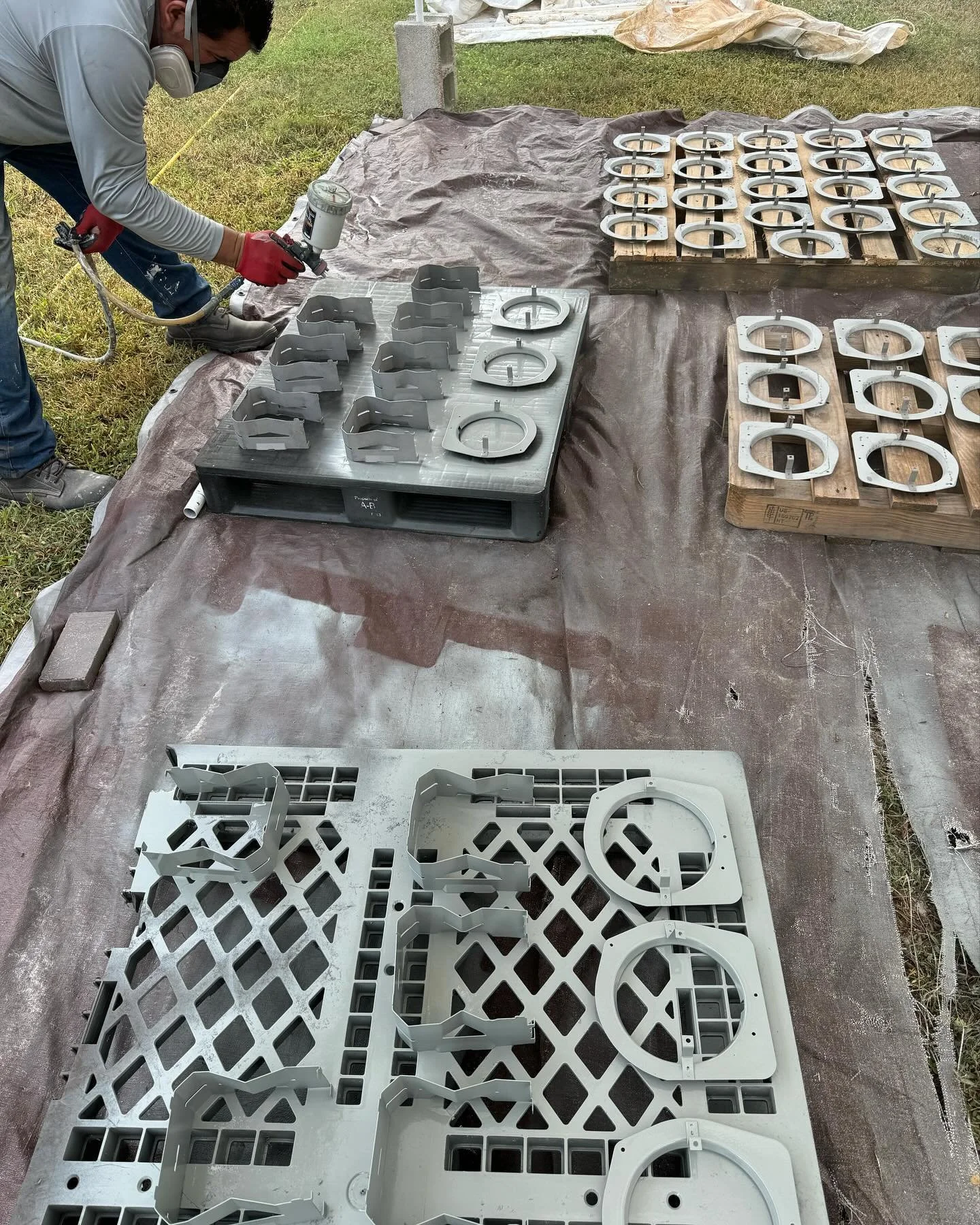 A person wearing gloves and a face mask is spray painting metal parts outdoors on a large tarp. The metal parts are arranged on pallets and a flat surface, likely for manufacturing or assembly.