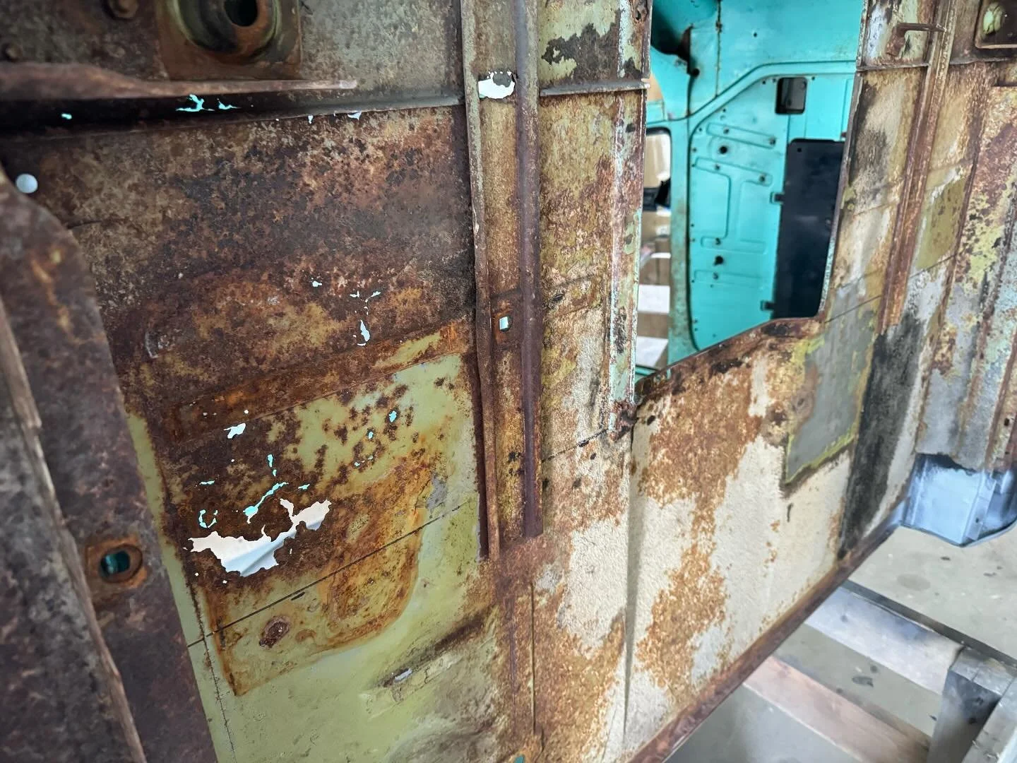 Close-up of a rusted, corroded metal surface with peeling paint, showing signs of wear and age, with part of a vintage vehicle interior visible in the background.