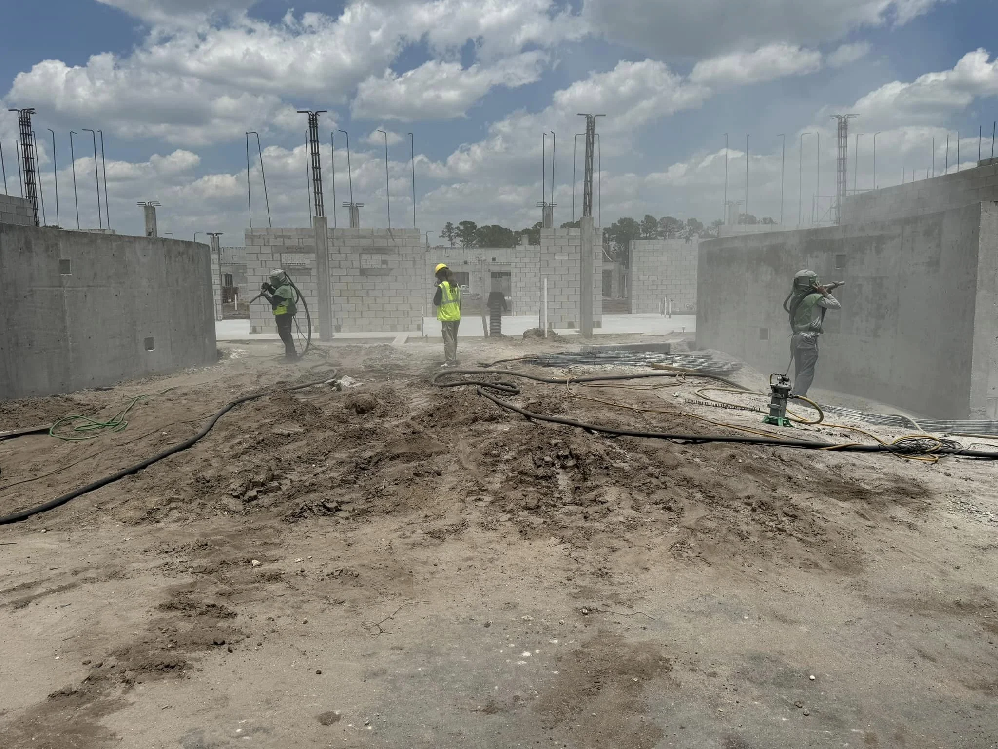 Construction workers wearing safety gear working on a construction site with unfinished brick and concrete structures, dirt ground, and tools, under a partly cloudy sky.
