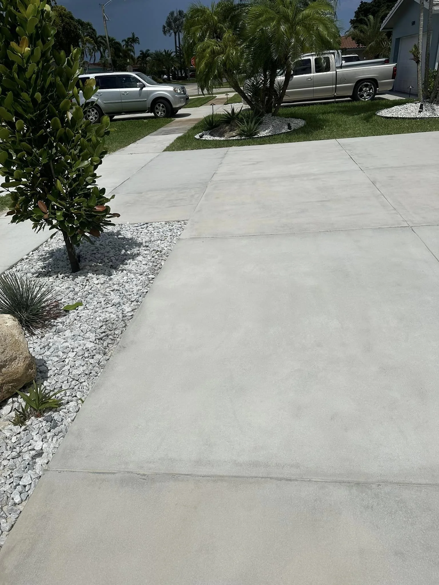 A sidewalk with decorative rocks and plants on the side, leading to a driveway with two parked trucks, with a grassy yard and palm trees in the background.