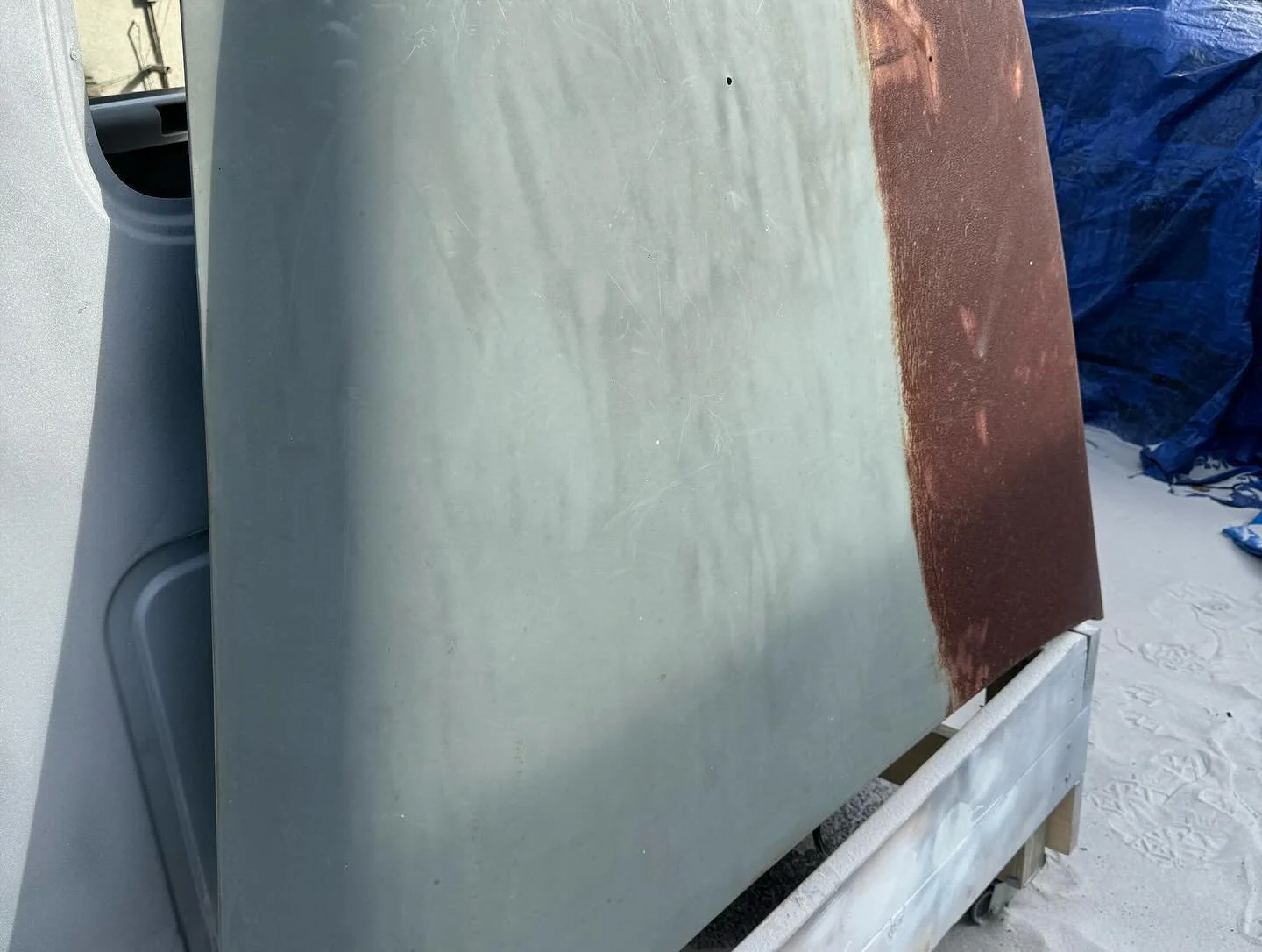 A metal surface with one half painted white and the other half rust-colored, sitting on a wooden frame outdoors.