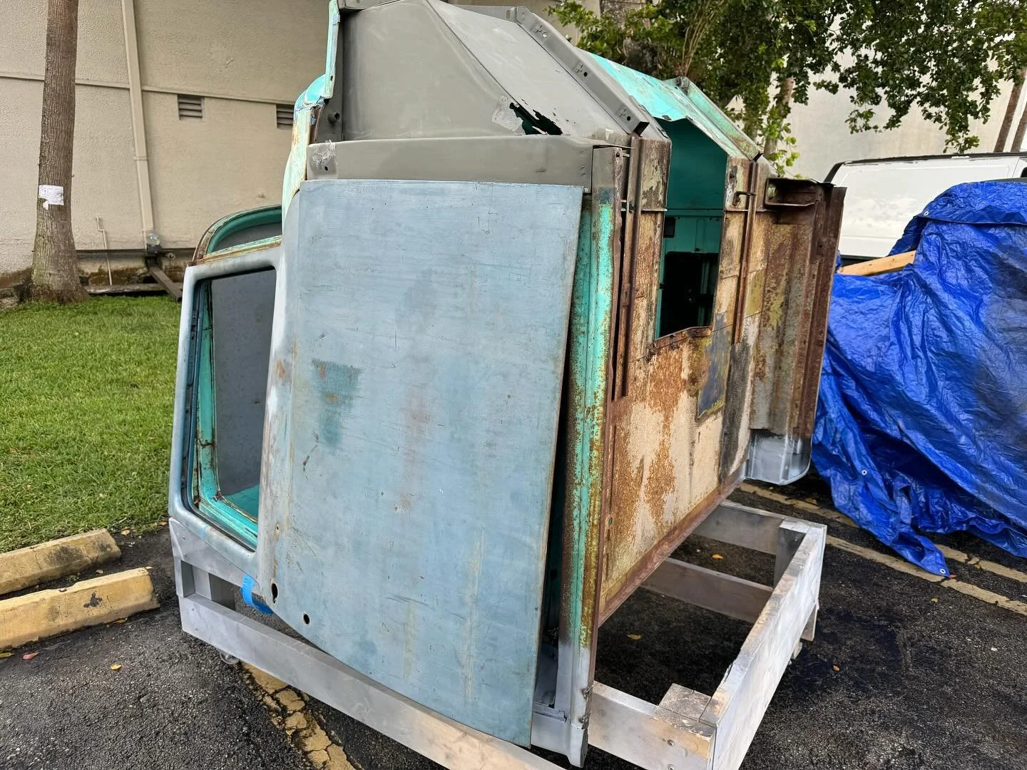 Old and rusty cash register or vending machine frame in an outdoor parking lot next to a blue tarp covering something.