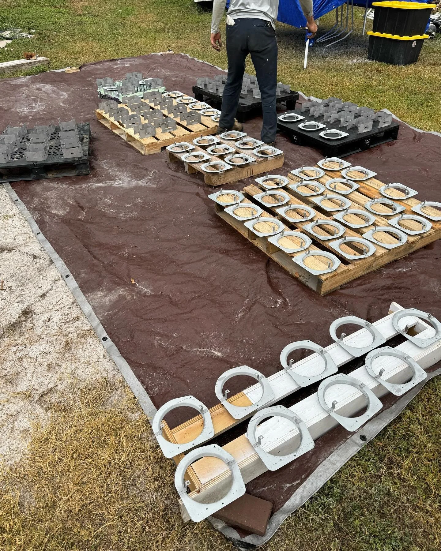 A person standing on a tarp-covered ground surrounded by hardware components, including large circular pieces, arranged on wooden pallets. The components appear to be parts for a construction or industrial project, with some mounted on black supports