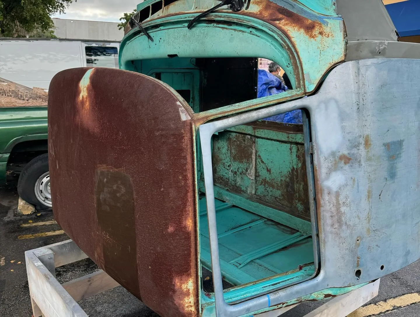 Rusty vintage truck cab with peeling turquoise and red paint, missing engine and interior, placed on a wooden frame outdoors.