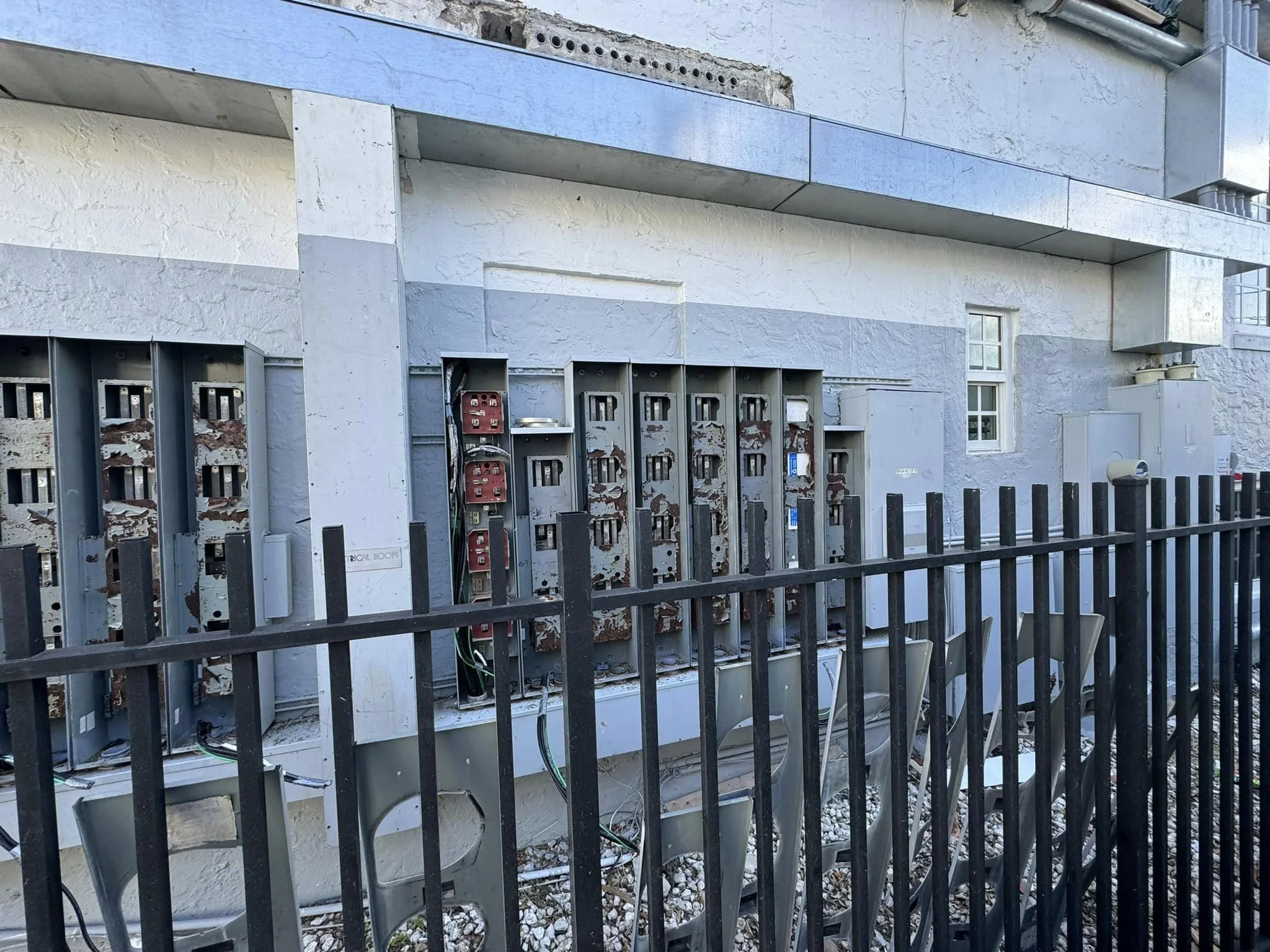 View of multiple electrical meters and panels attached to the exterior wall of a building, behind a black metal fence.