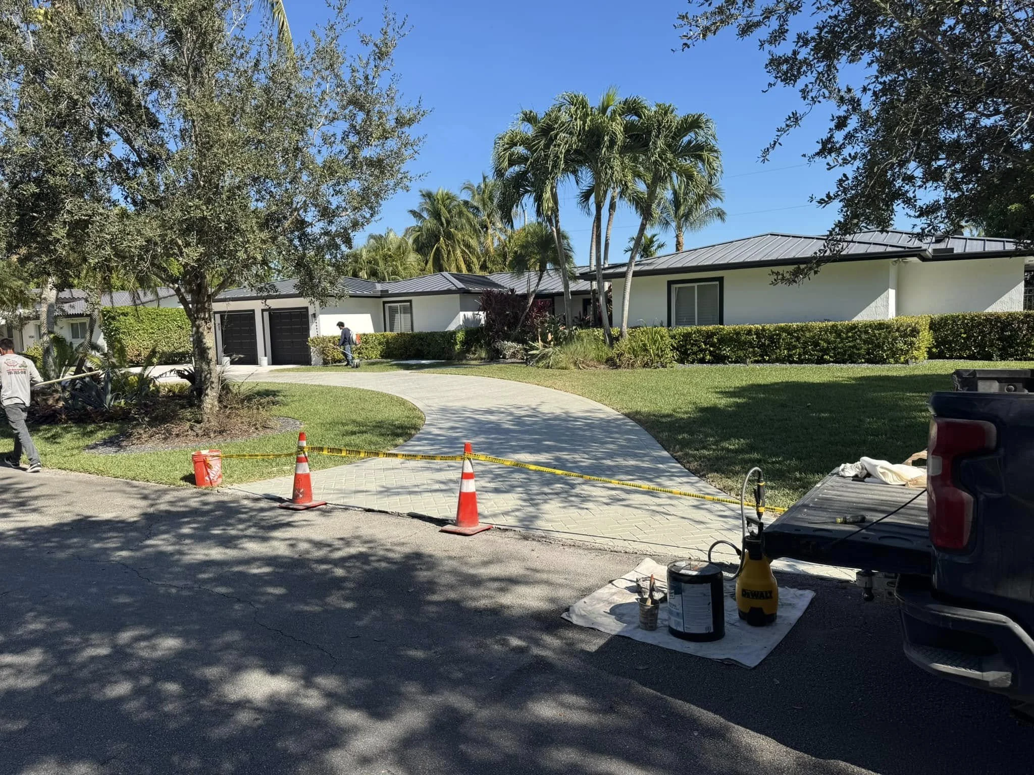 A curved concrete sidewalk leading to a white residential house with a metal roof, surrounded by palm trees and greenery. Construction work is in progress with orange cones, caution tape, and tools near the sidewalk.