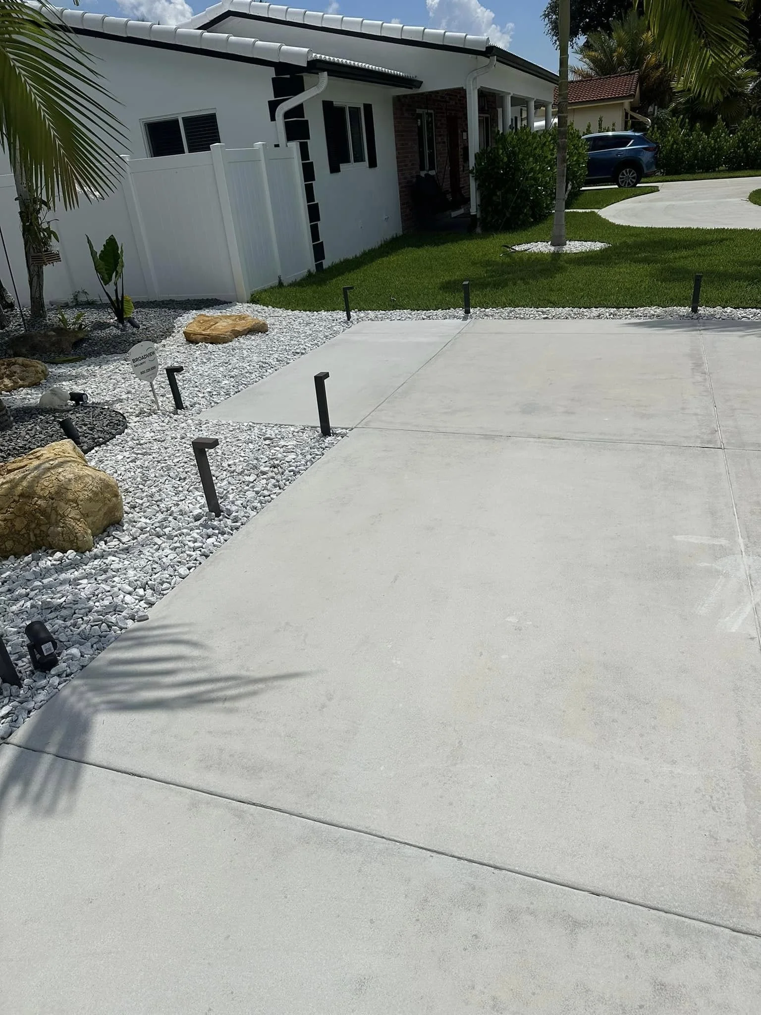 Concrete driveway in front of a house with landscaping, rocks, plants, small fence, green lawn, trees, and a parked car.