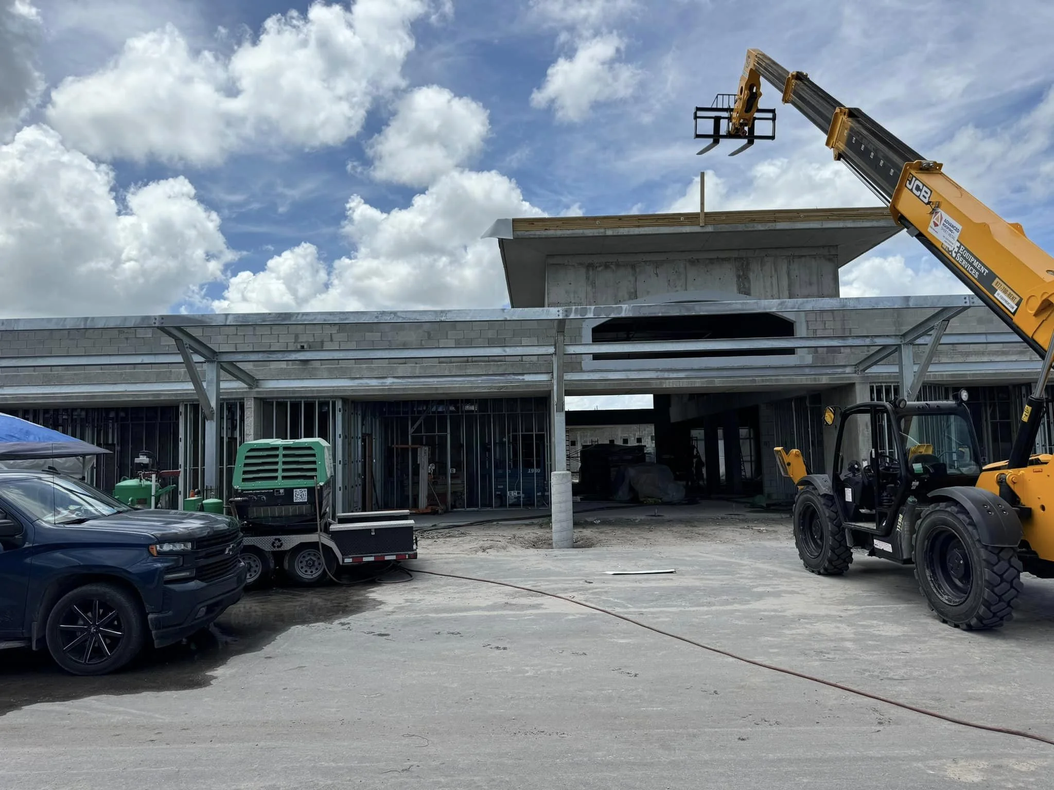 Construction site with an unfinished building, a forklift, and a parking lot with vehicles under a partly cloudy sky.