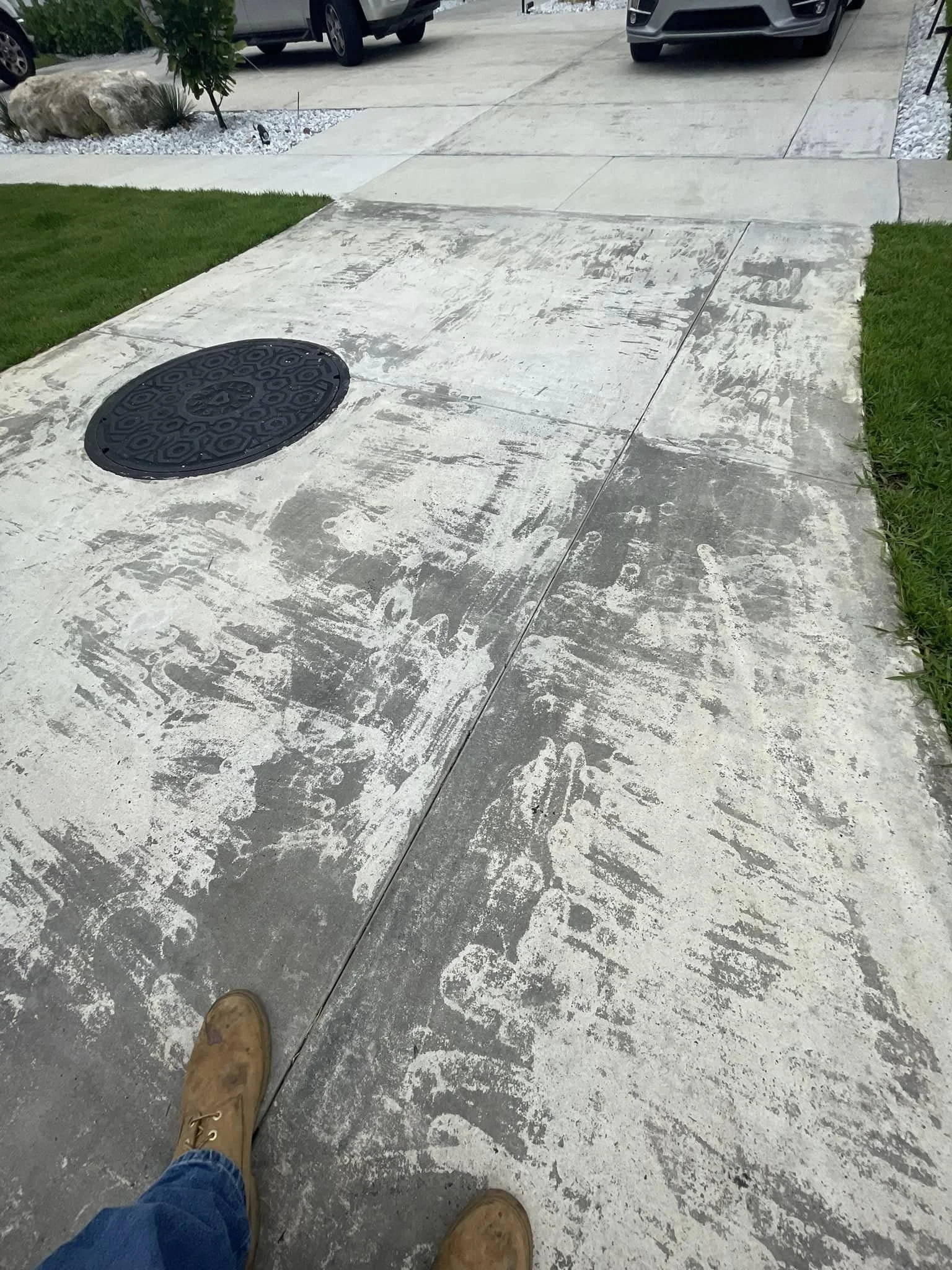 View of a concrete sidewalk with a black circular utility cover, a portion of brown work boots and blue jeans, and a partial view of a driveway with parked cars in the background.