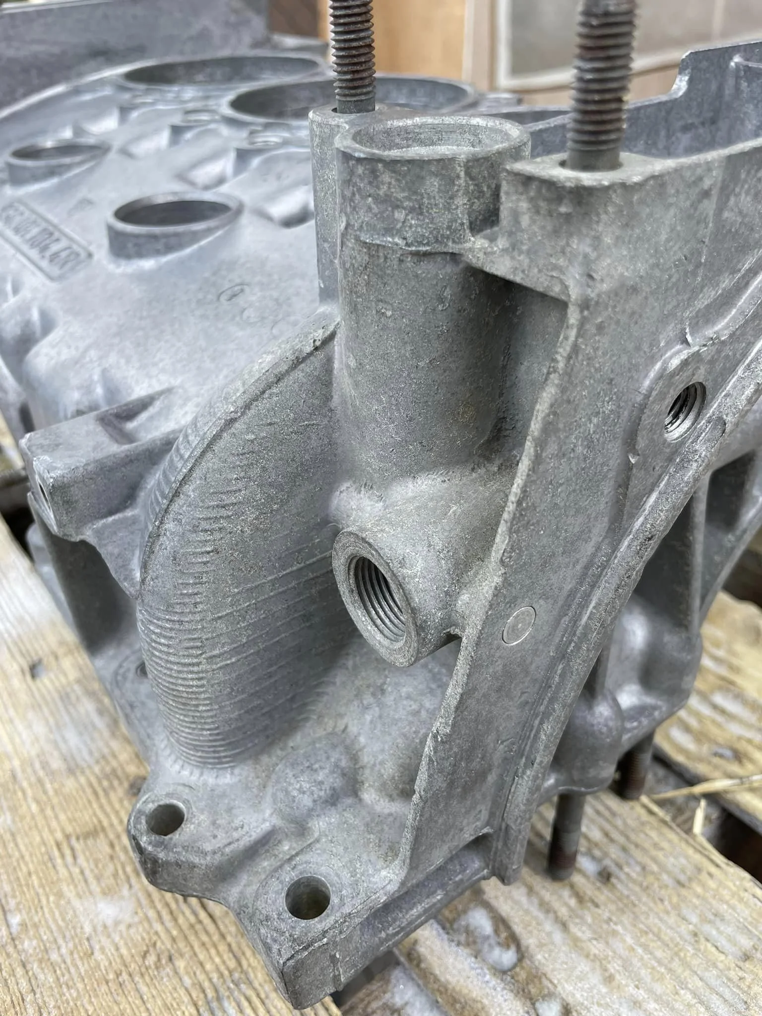 Close-up of a metallic engine block with threaded holes and mounting points, placed on a wooden surface.