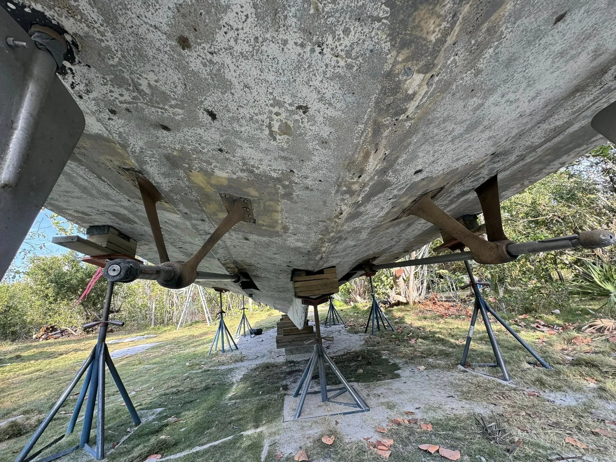 Underside of a boat supported by multiple jack stands on land, showing weathered and peeling paint with trees and grass in the background.