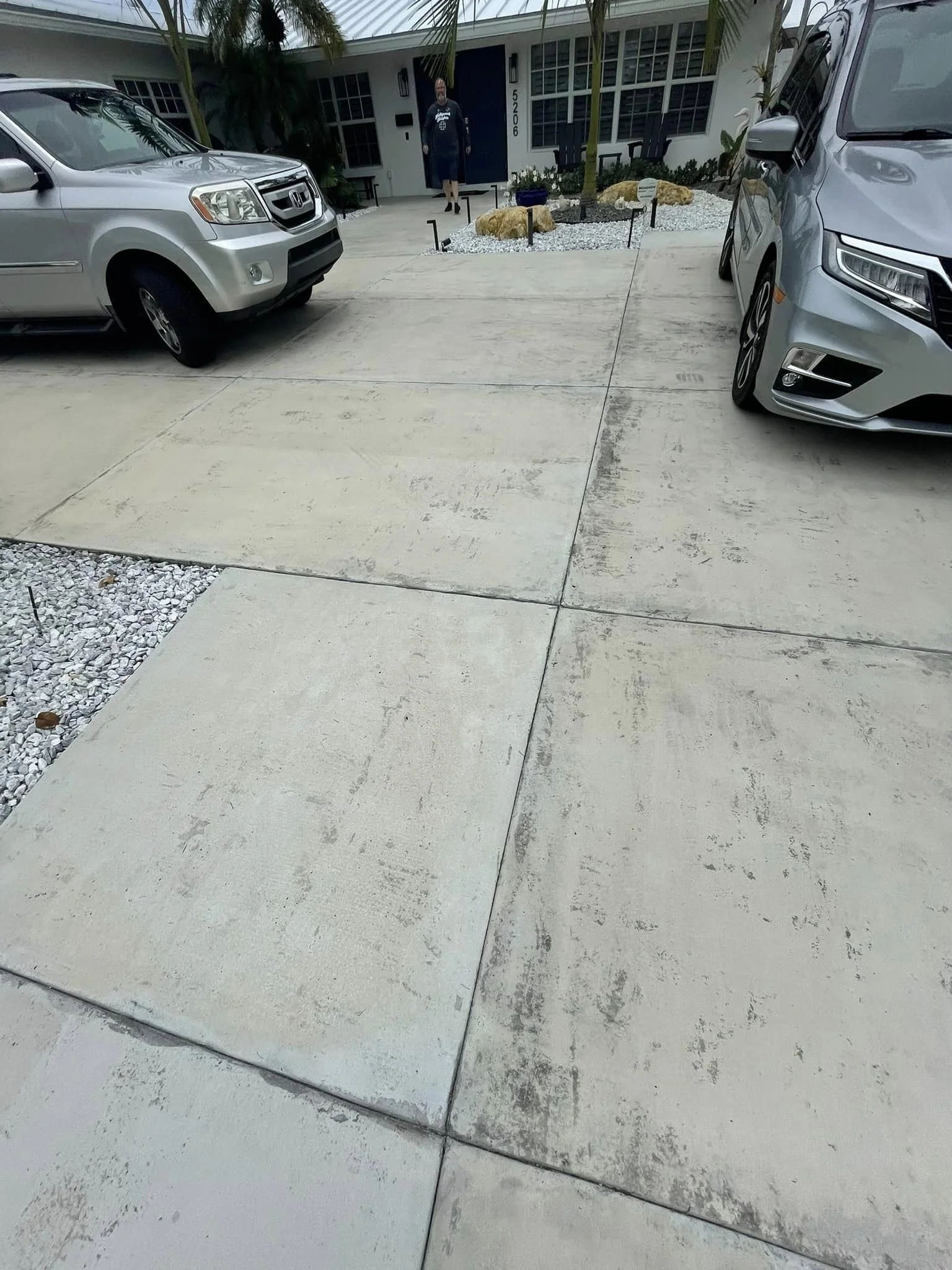 Concrete driveway in front of a house with two parked cars, one on each side, and a person standing in front of the house.