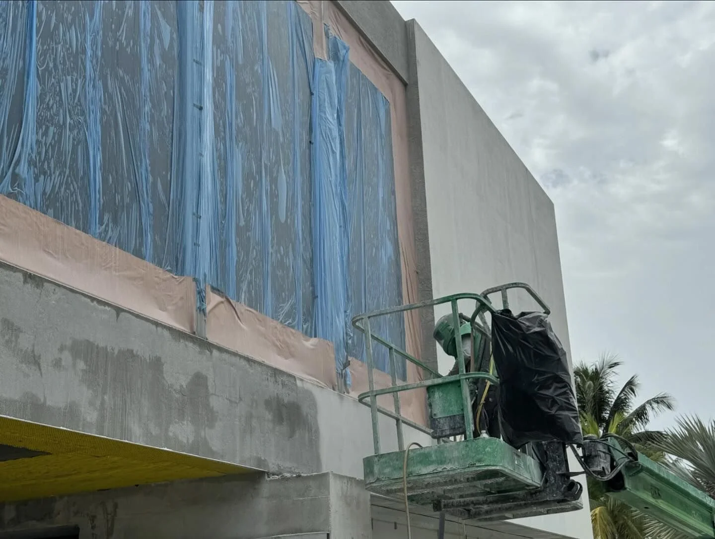 Construction worker on a green lift working on the exterior wall of a building, with part of the wall covered in blue plastic sheeting and beige painter's tape, under a partly cloudy sky.