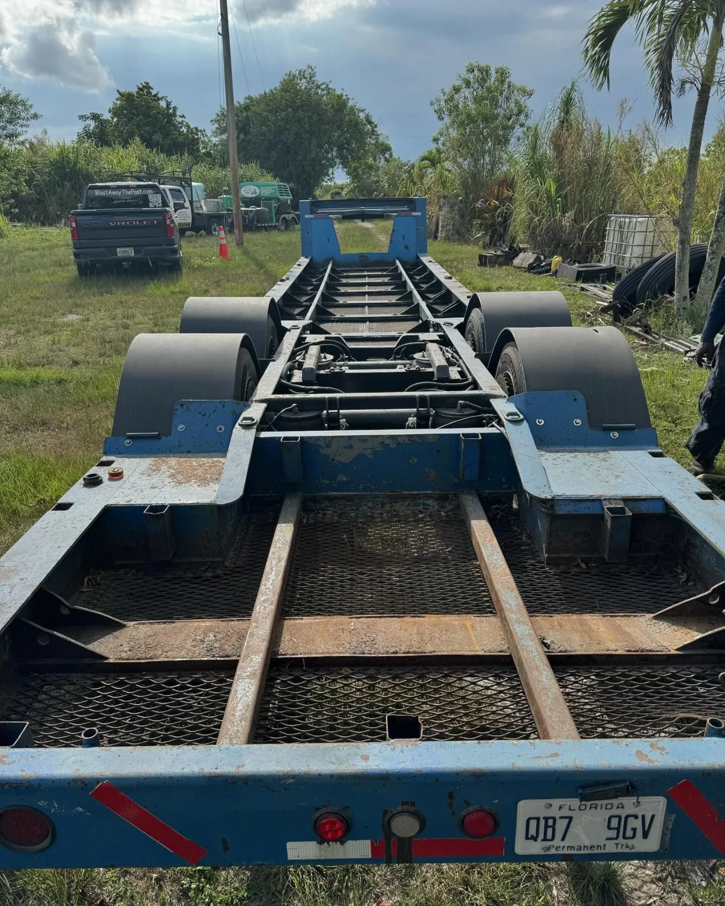 View of a rusty blue trailer chassis with large black tires on a grassy outdoor area, with vehicles and trees in the background. getting ready for sandblasting