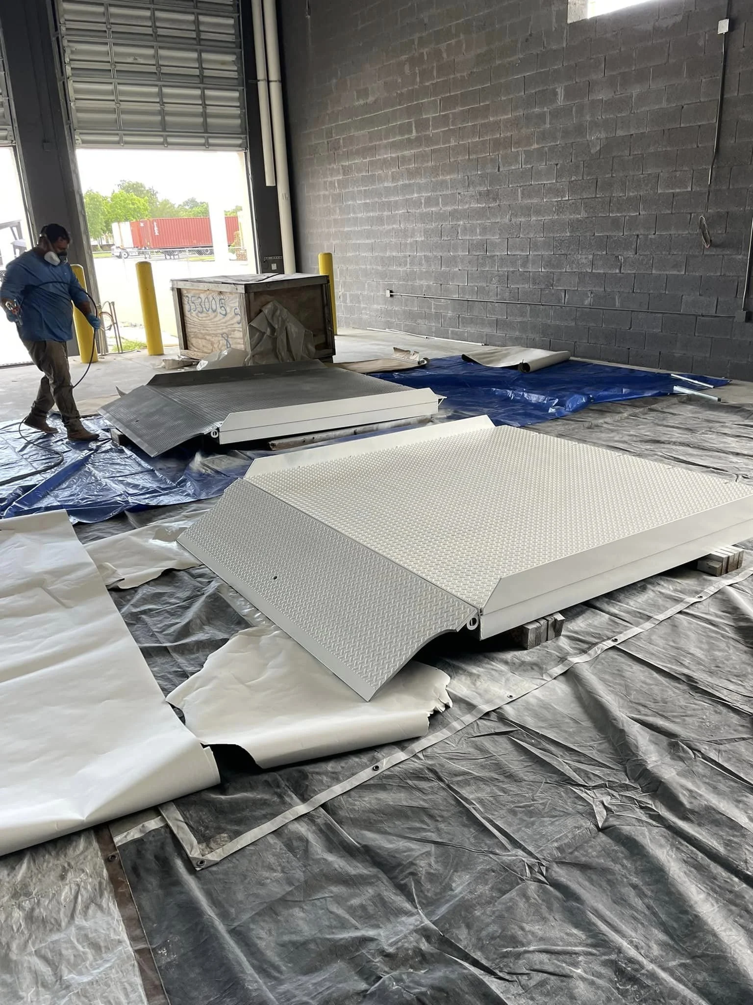 Worker in blue jacket preparing metal platform and ramp inside warehouse with open garage door, showing parked trucks outside.