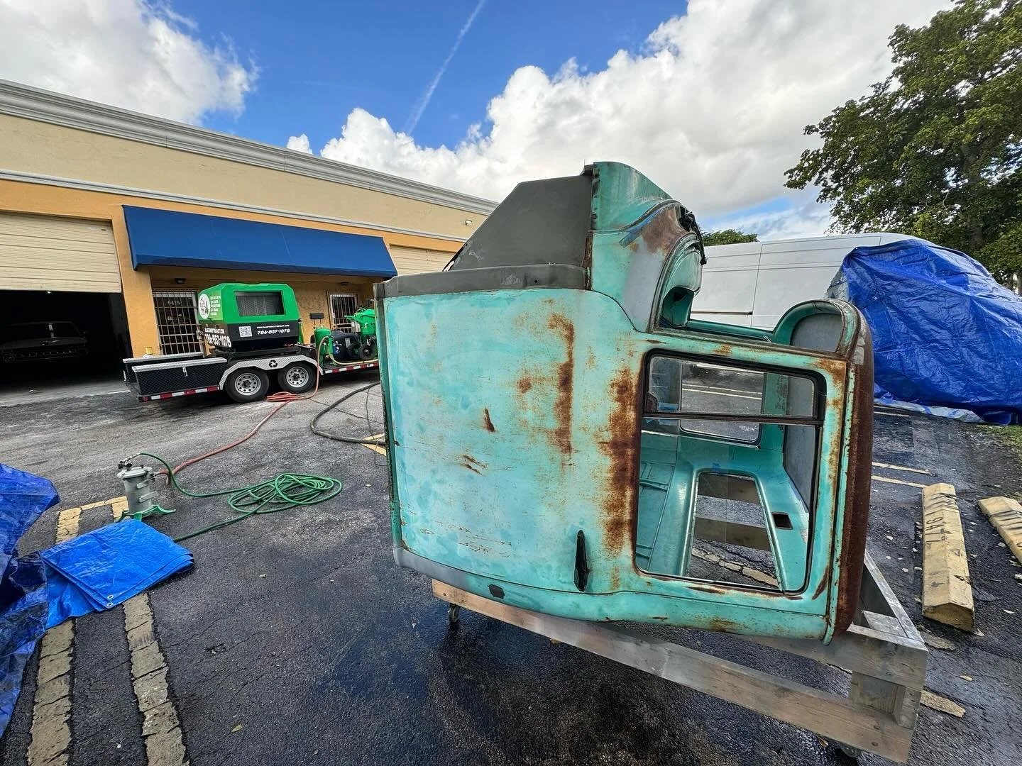 Old, rusty, turquoise-colored vehicle cabin, possibly from a truck, placed on a wooden platform outdoors in a parking lot, with tarp-covered objects and a building in the background.