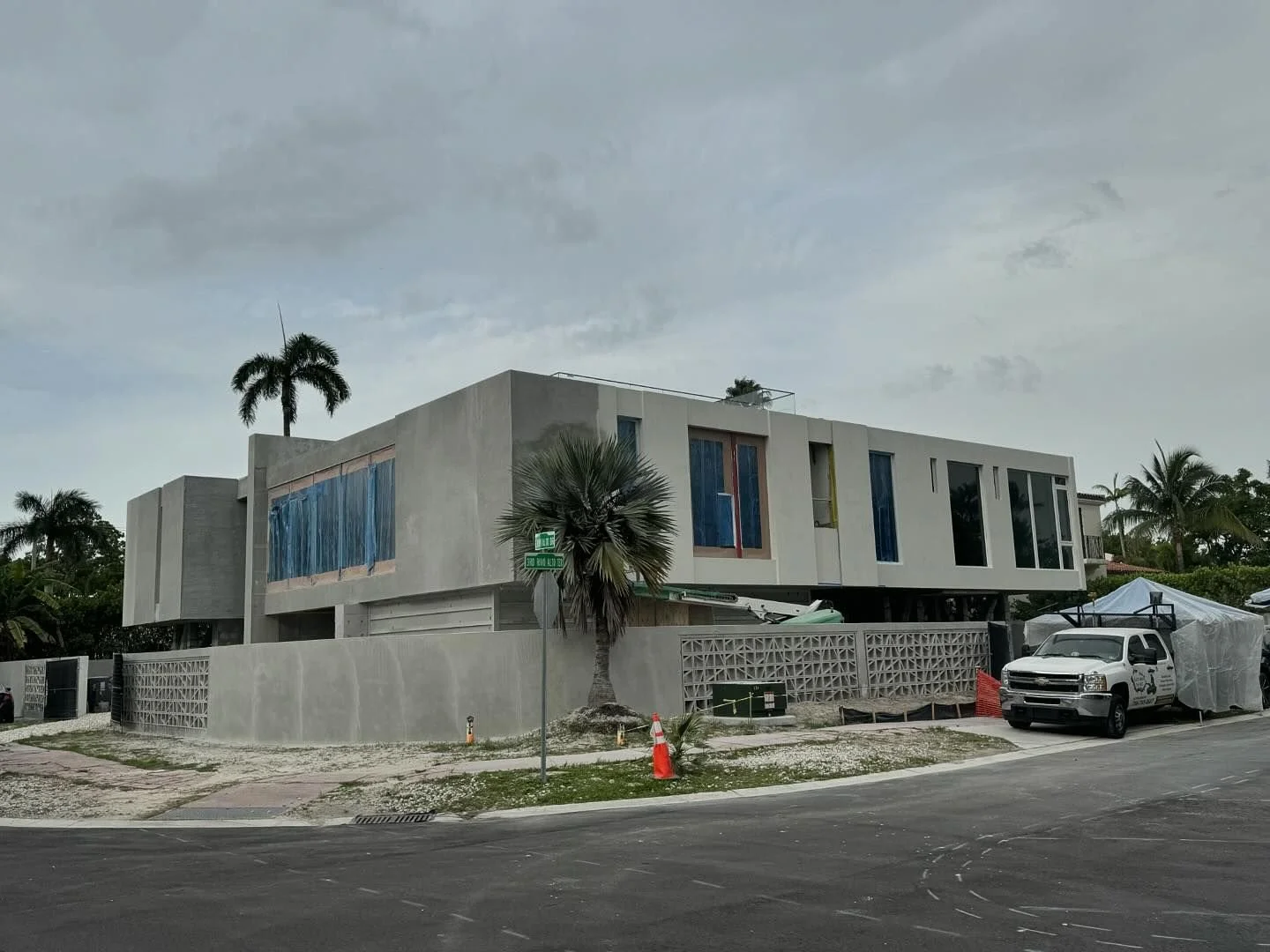 A modern, multi-story house under construction with large windows, palm trees, and construction vehicles parked outside on a cloudy day.