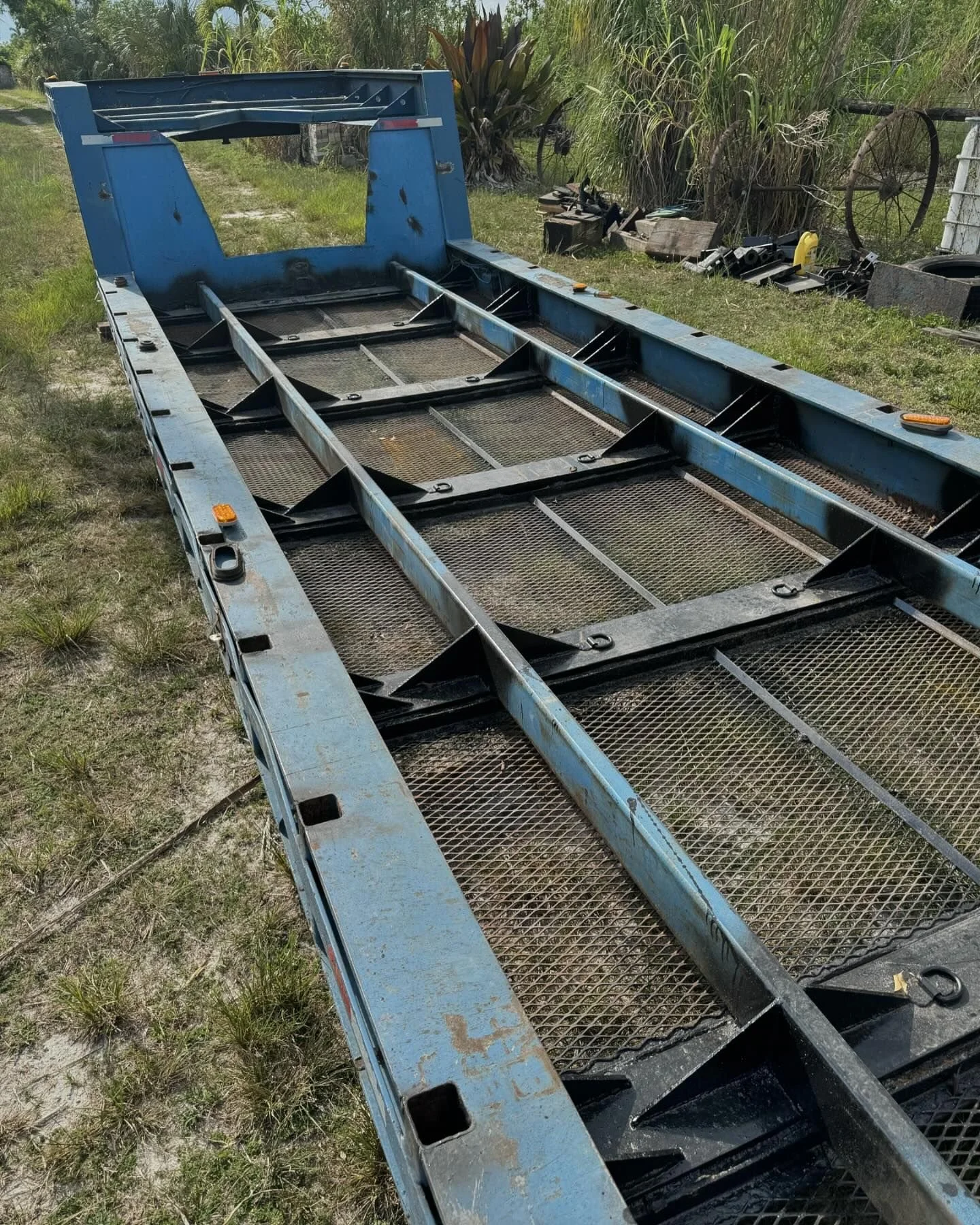 View of a disassembled blue trailer with metal mesh flooring lying on grass with some tools and machinery parts in the background.