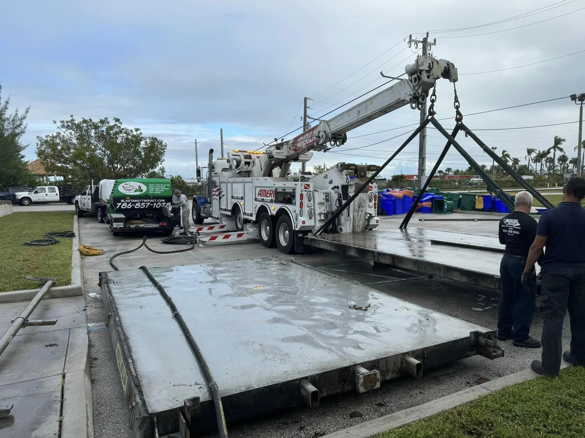 Utility workers are using a large crane truck and flatbed to lift and transport a large metal structure, with hoses and cables on the ground, while two men discuss nearby in an outdoor parking lot.