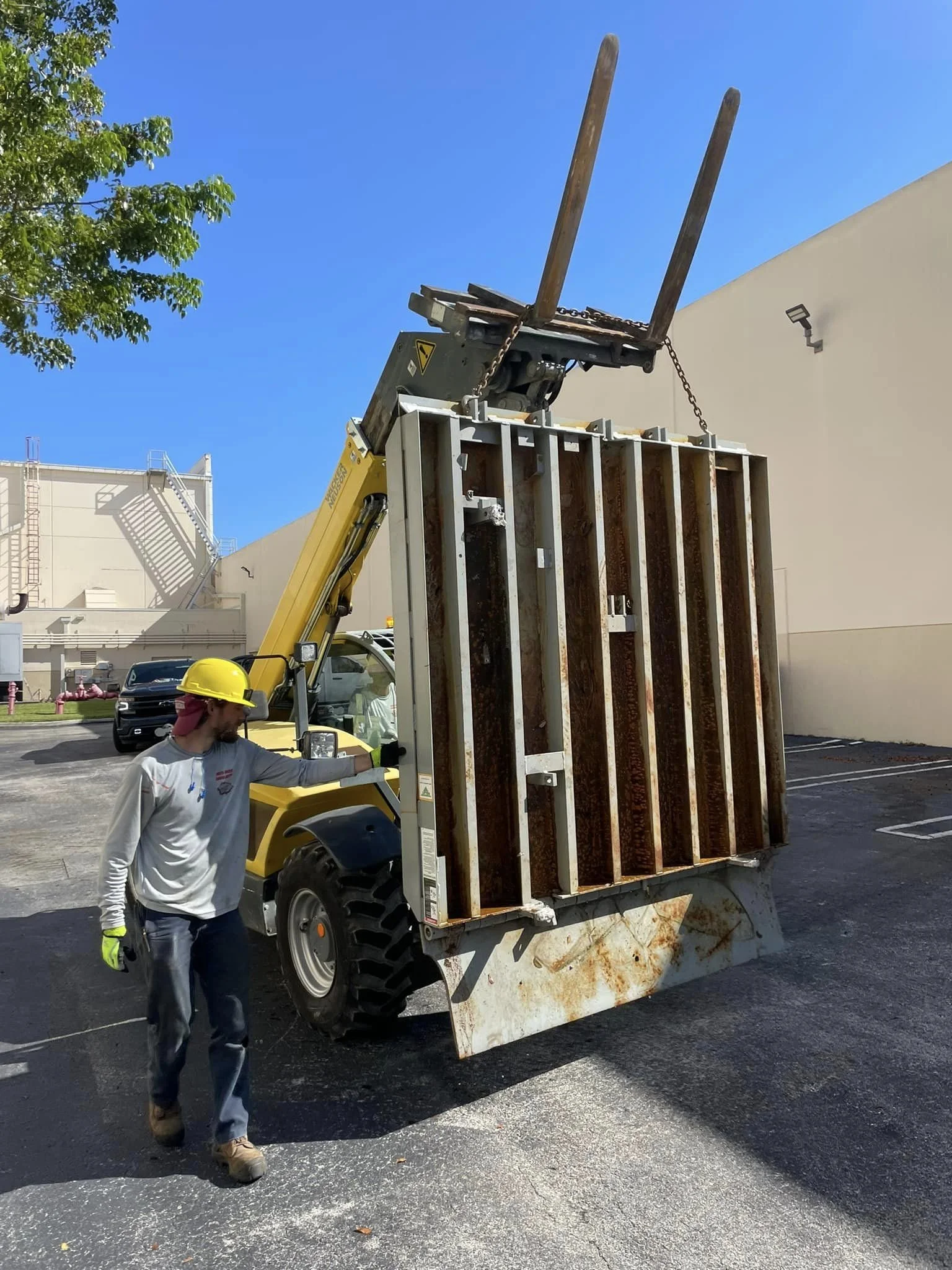 Worker in a yellow safety helmet and gloves checks a large, rusty, metal container attached to a yellow forklift in a parking lot under a blue sky.