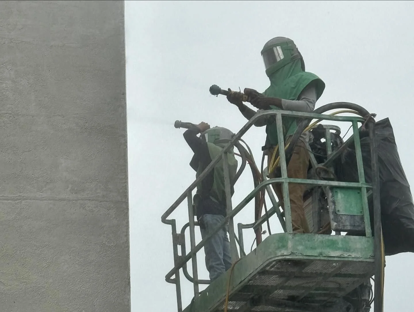 Two construction workers on a green elevated work platform, cleaning a building's exterior wall with pressure washers, wearing protective gear including helmets and face shields.