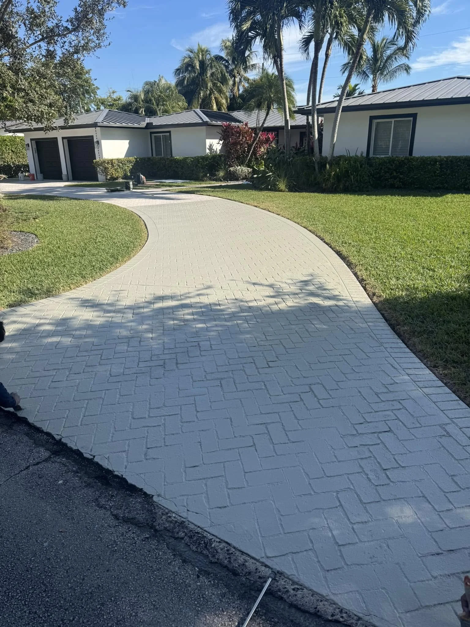 A curved, light-colored brick driveway leading to a house with white exterior, dark garage doors, and tropical landscaping, including palm trees and lush bushes.