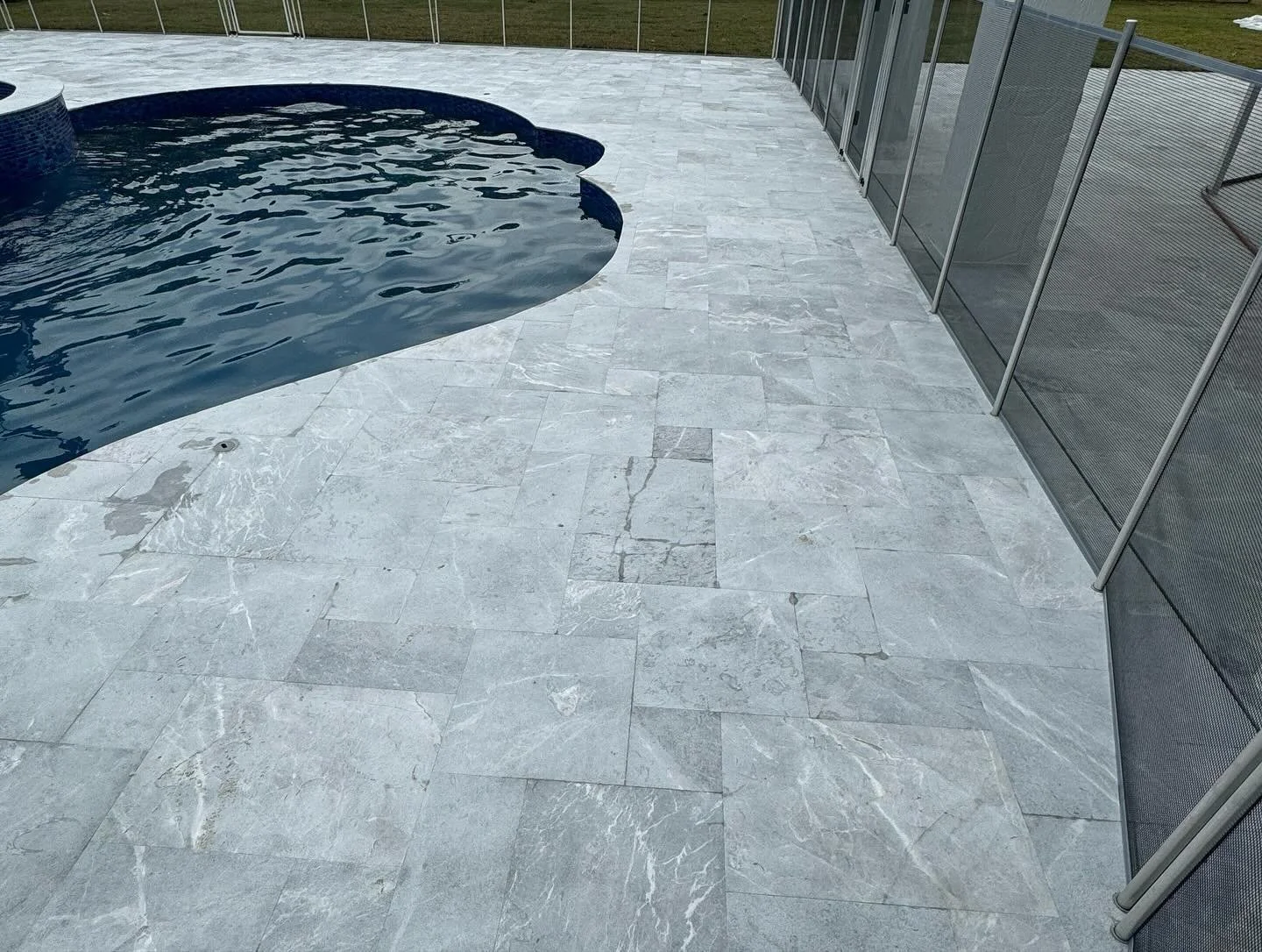 Patio with light gray stone tiles adjacent to a swimming pool with light dark blue water, enclosed by a black metal fence.