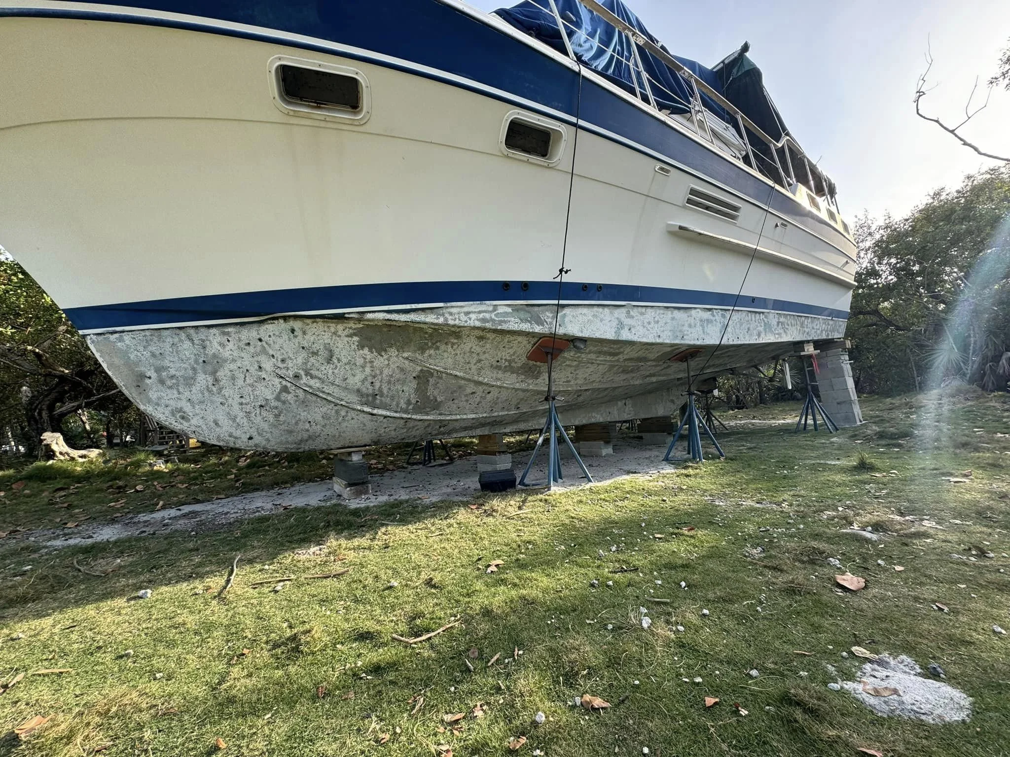 A boat on land raised on stands and blocks, showing its underside which is stained and dirty, with trees and grass in the background.
