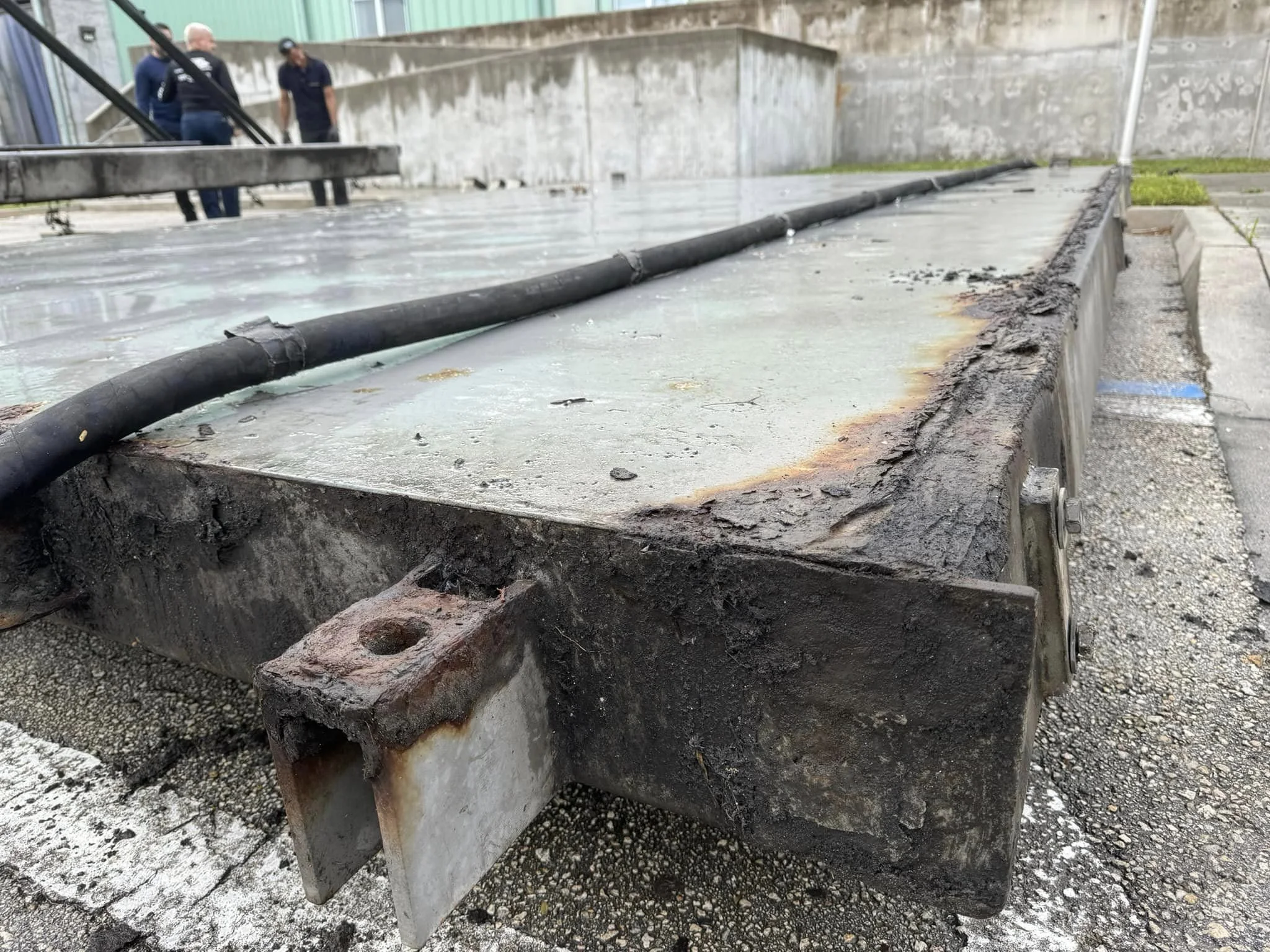 Close-up of a large industrial metal tank or container with a black pipe on top, showing signs of rust and dirt, outdoors near a concrete wall with two people in the background.