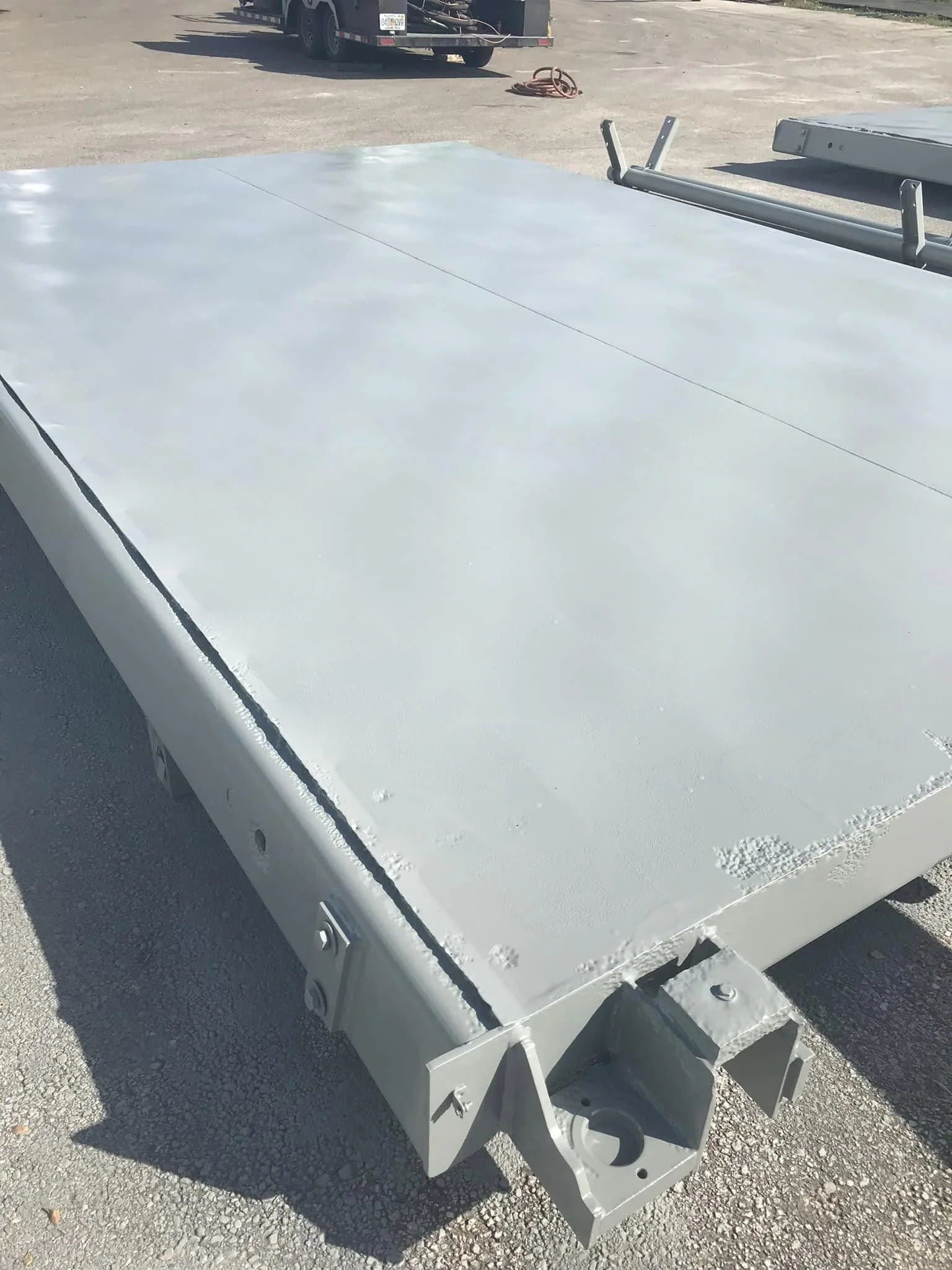 A large, flat, gray metal surface of a trailer or platform with some hardware attachment points, outdoors on pavement.