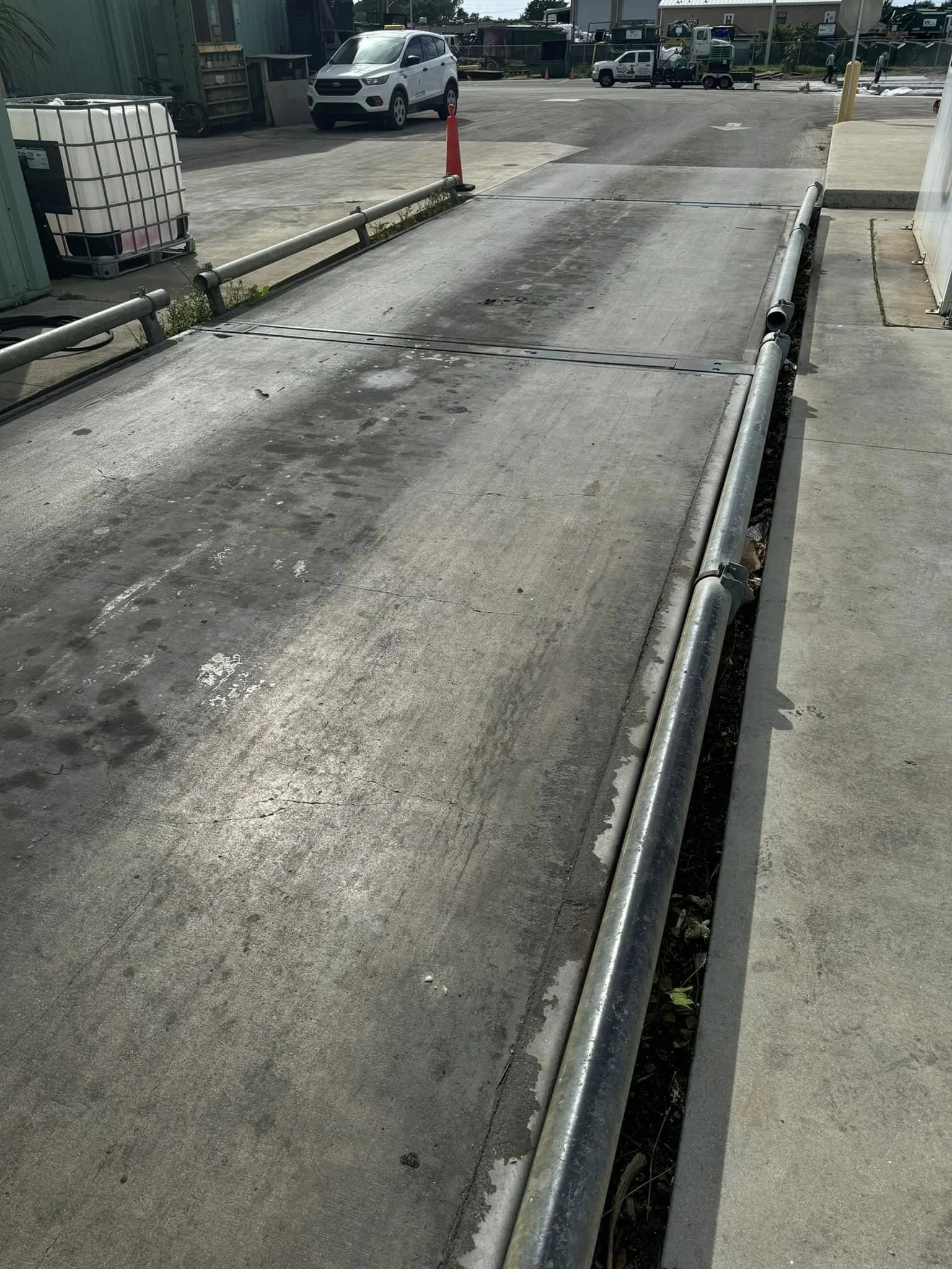 Concrete driveway with rubber safety barriers and metal pipes along the edges, leading to a parking area with vehicles and construction equipment in the background.