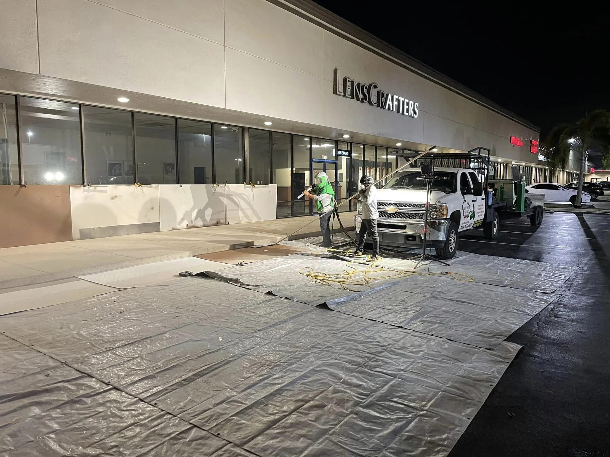 Workers cleaning outside a store named LensCrafters at night, using hoses and equipment on large sheets spread on the ground in a parking lot.