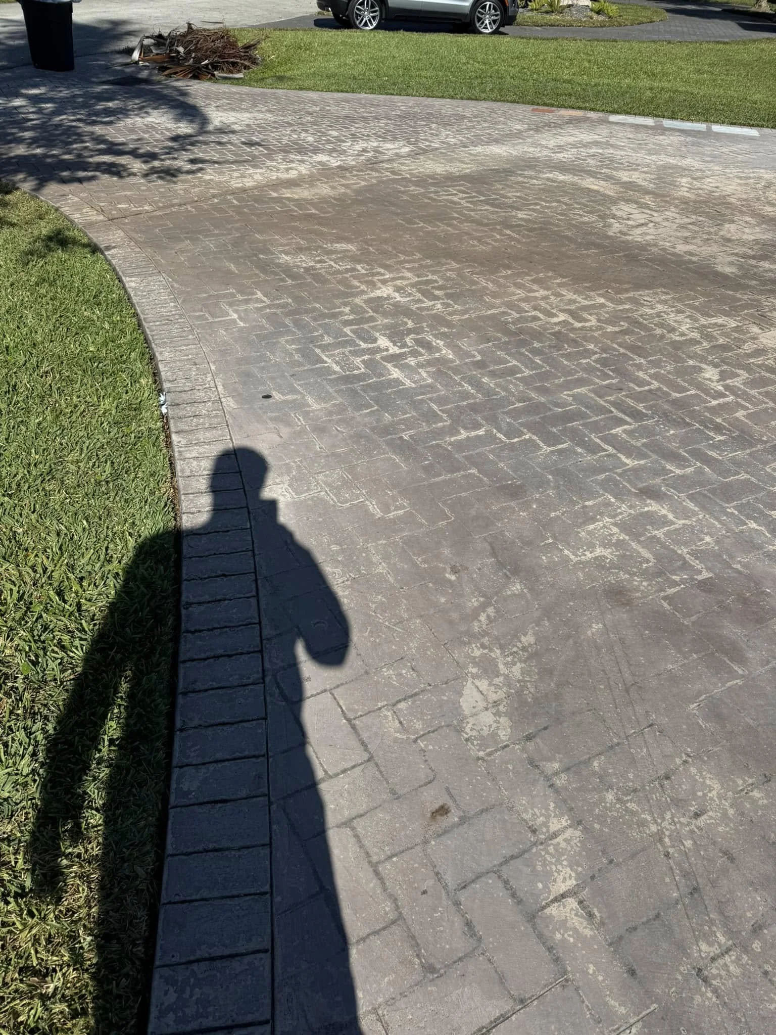 Shadow of a person holding a camera cast on a paved walkway and grass, with parked cars and bushes in the background.