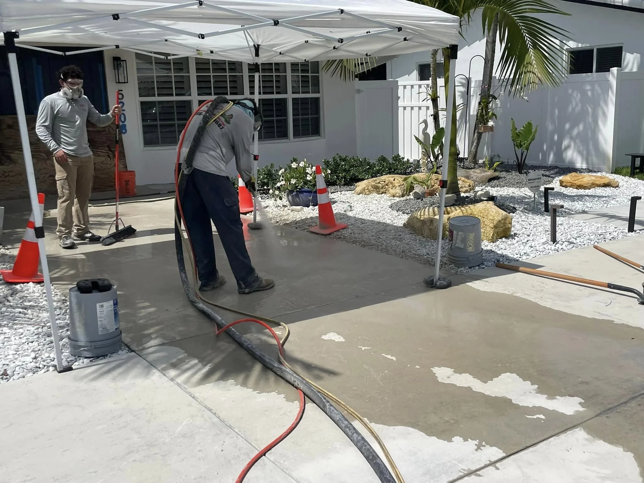 Workers power-washing a concrete sidewalk in a residential yard with a white canopy tent, white gravel, rock landscaping, and palm trees.