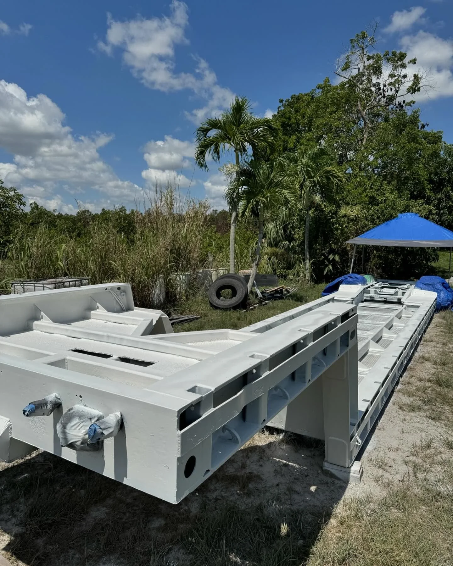Large white boat hull lying on the ground outdoors with trees, grass, and a blue sky with clouds in the background. There is a blue canopy tent and a tire nearby.